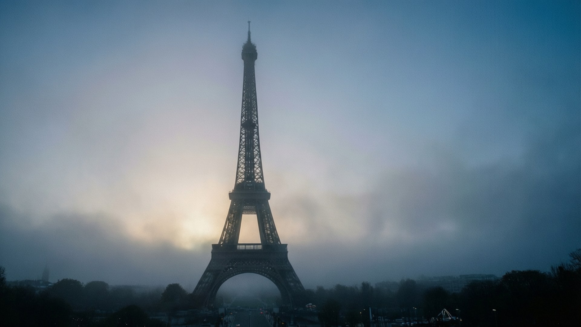Misty Morning Eiffel 壁紙 — 艾菲爾鐵塔在霧濛濛的黎明天空下，以黑暗的輪廓矗立，柔和的金色光芒穿透雲層。