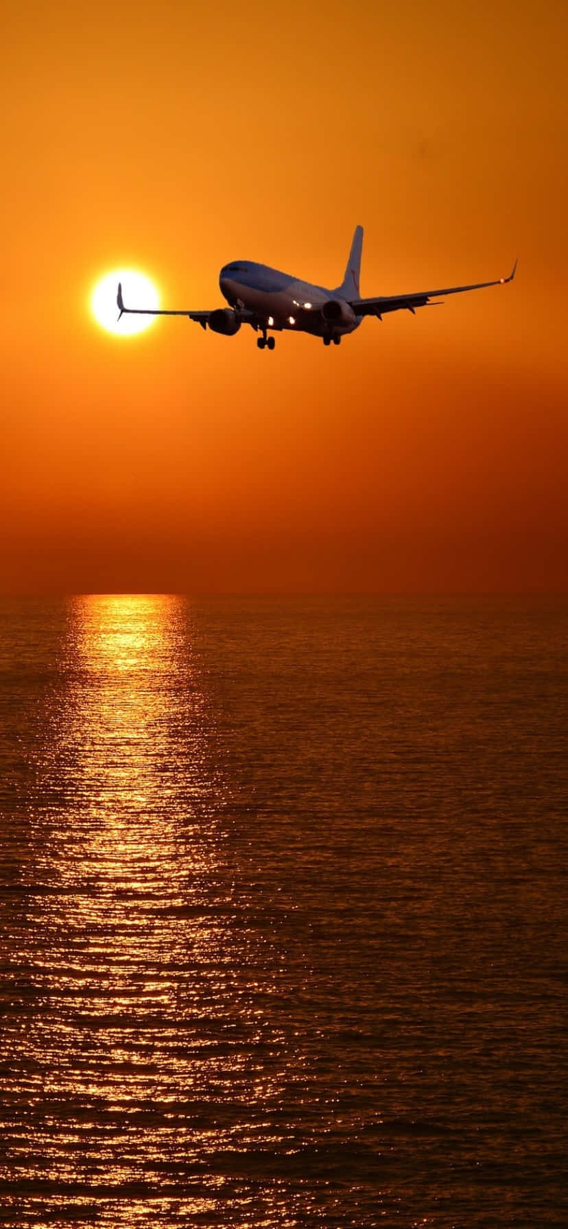  壁紙 — 一架飛機在閃閃發光的海洋上空低飛，以發光的橙色日落為背景形成剪影。