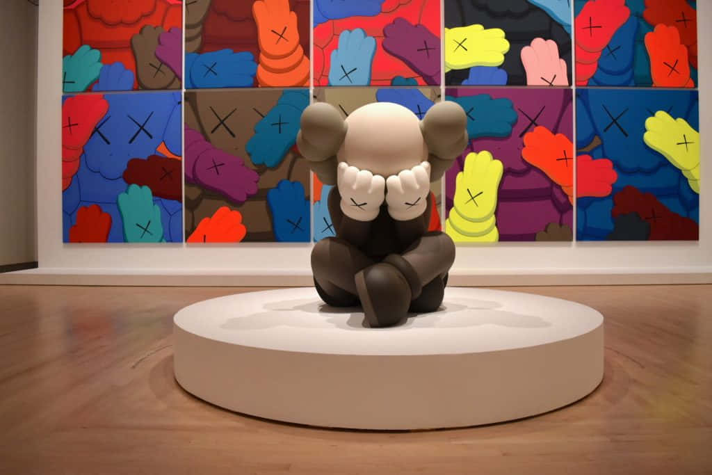 KAWS 桌布 壁紙 — AKAWSleepy 雕塑坐在白色底座上，前面是畫廊中色彩繽紛的 KAWS 藝術品網格。