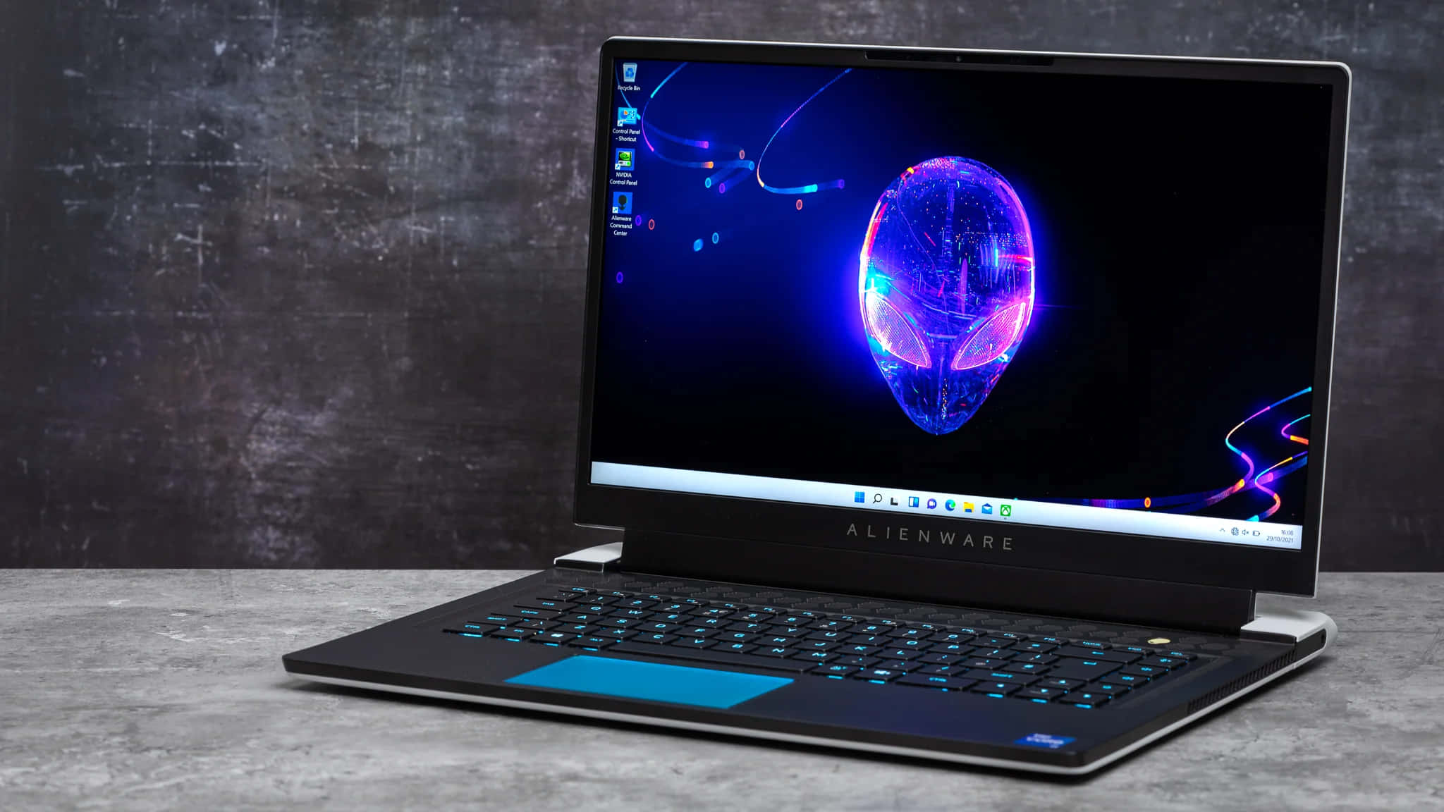 外星人 桌布 壁紙 — Alienware 筆記型電腦帶有發光外星人頭像桌布，放在混凝土上，背景為深色紋理牆壁。