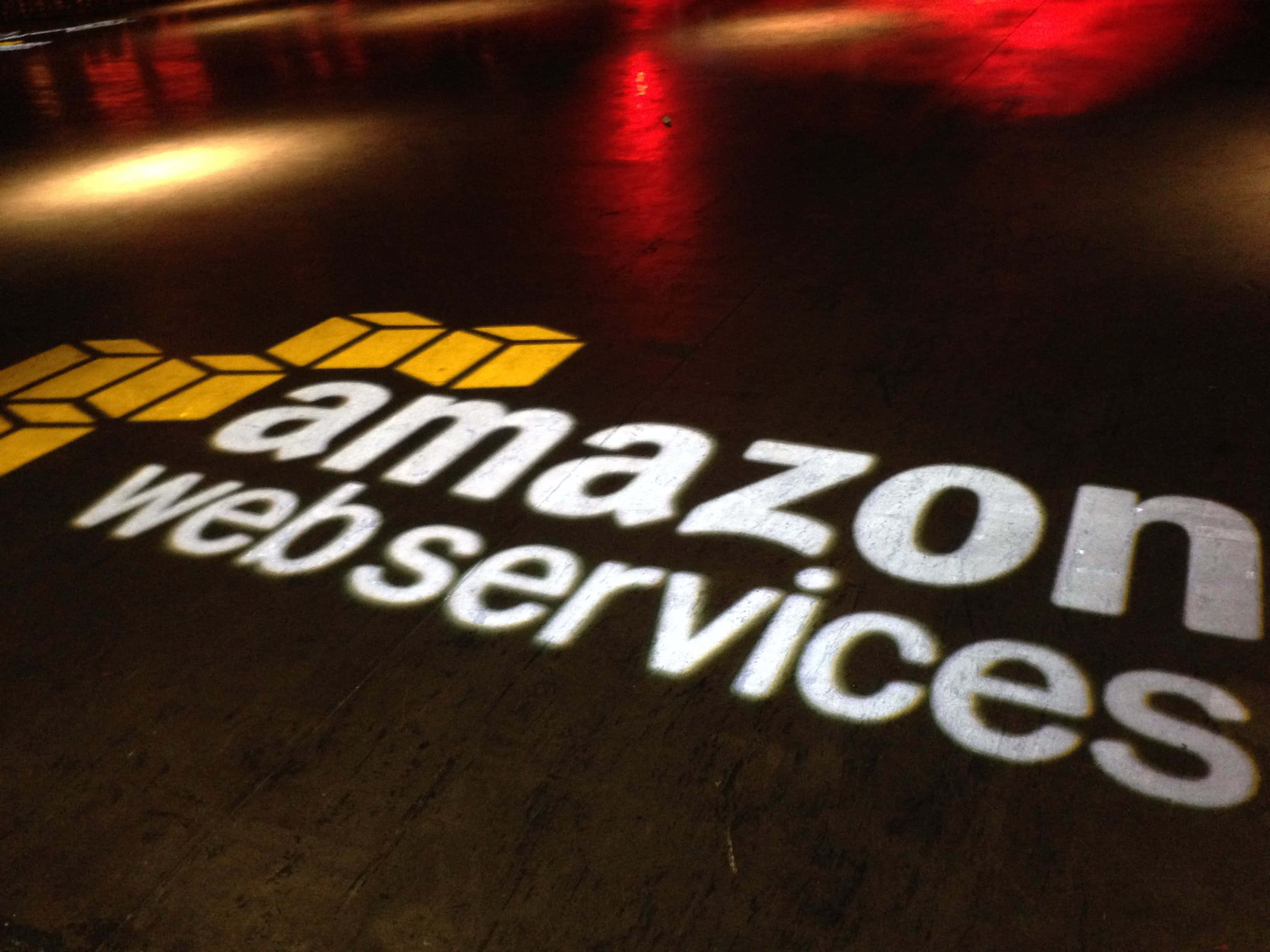 亞馬遜 桌布 壁紙 — Amazon Web Services 標誌以白色和黃色光線投射在深色紋理地板上，帶有紅色和黃色的環境反射。
