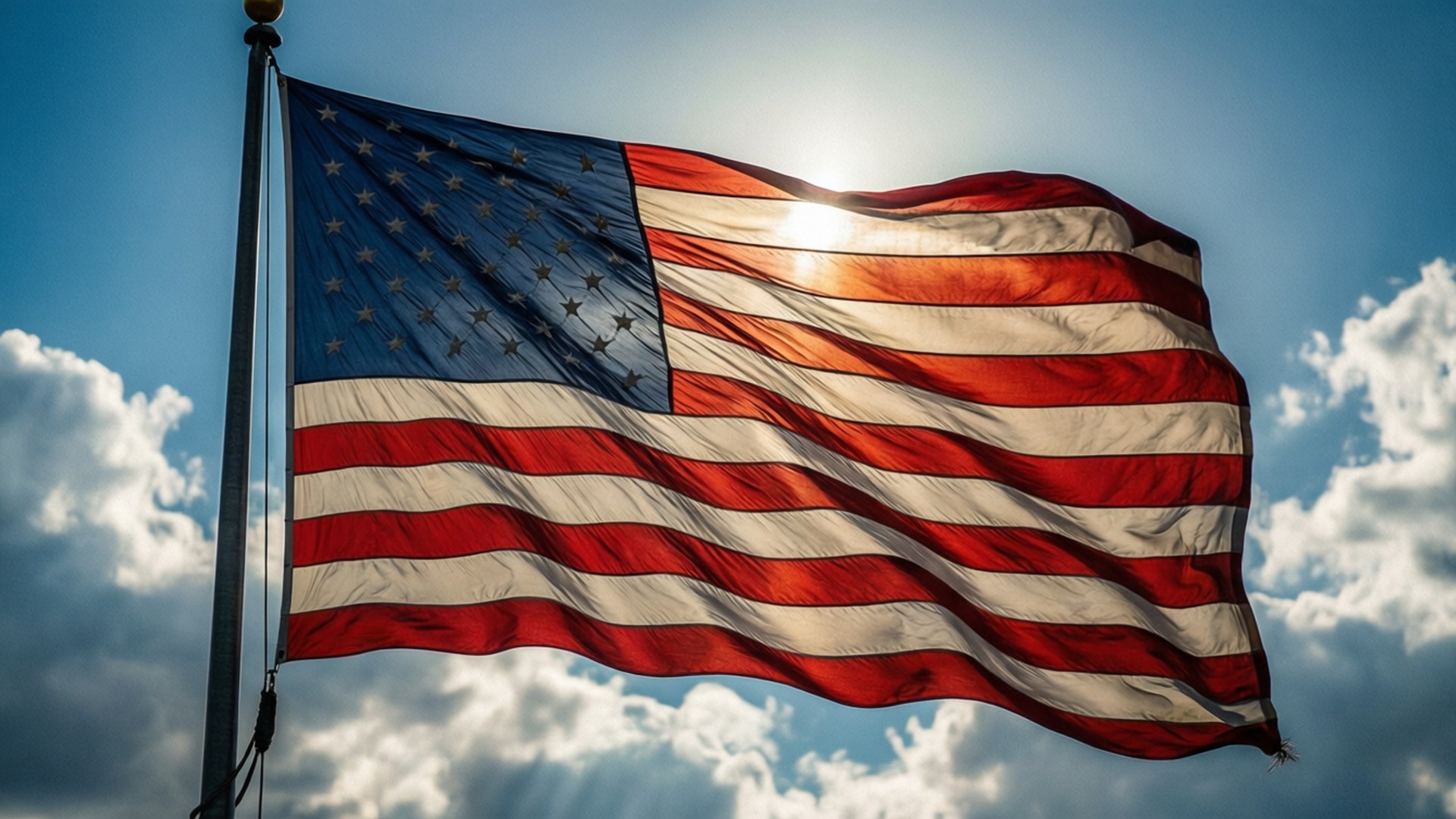 Stars and Stripes Sky 壁紙 — 美國國旗在明亮的藍天、蓬鬆的白雲和陽光穿透條紋的背景下自豪地飄揚。