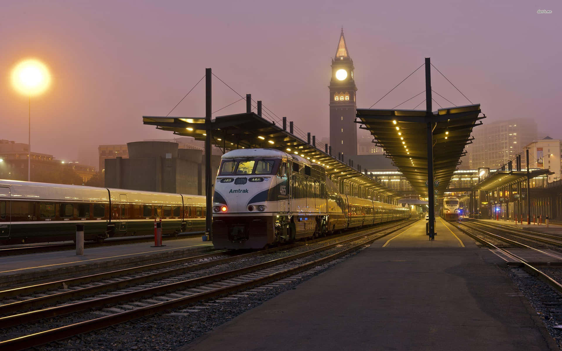  壁紙 — Amtrak 列車在黃昏時分滑過煙霧瀰漫的車站，背景中鐘樓閃爍著光芒。