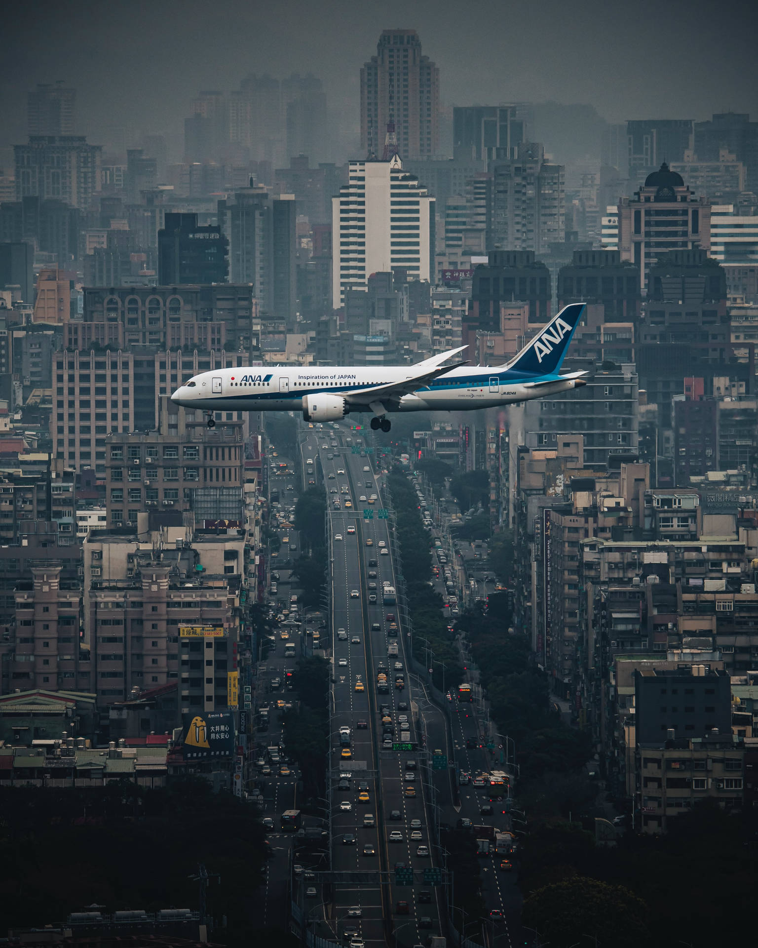  壁紙 — 一架全日空飛機在灰濛濛的天空下，低飛越過密集的城市天際線，下方是繁忙的高速公路。