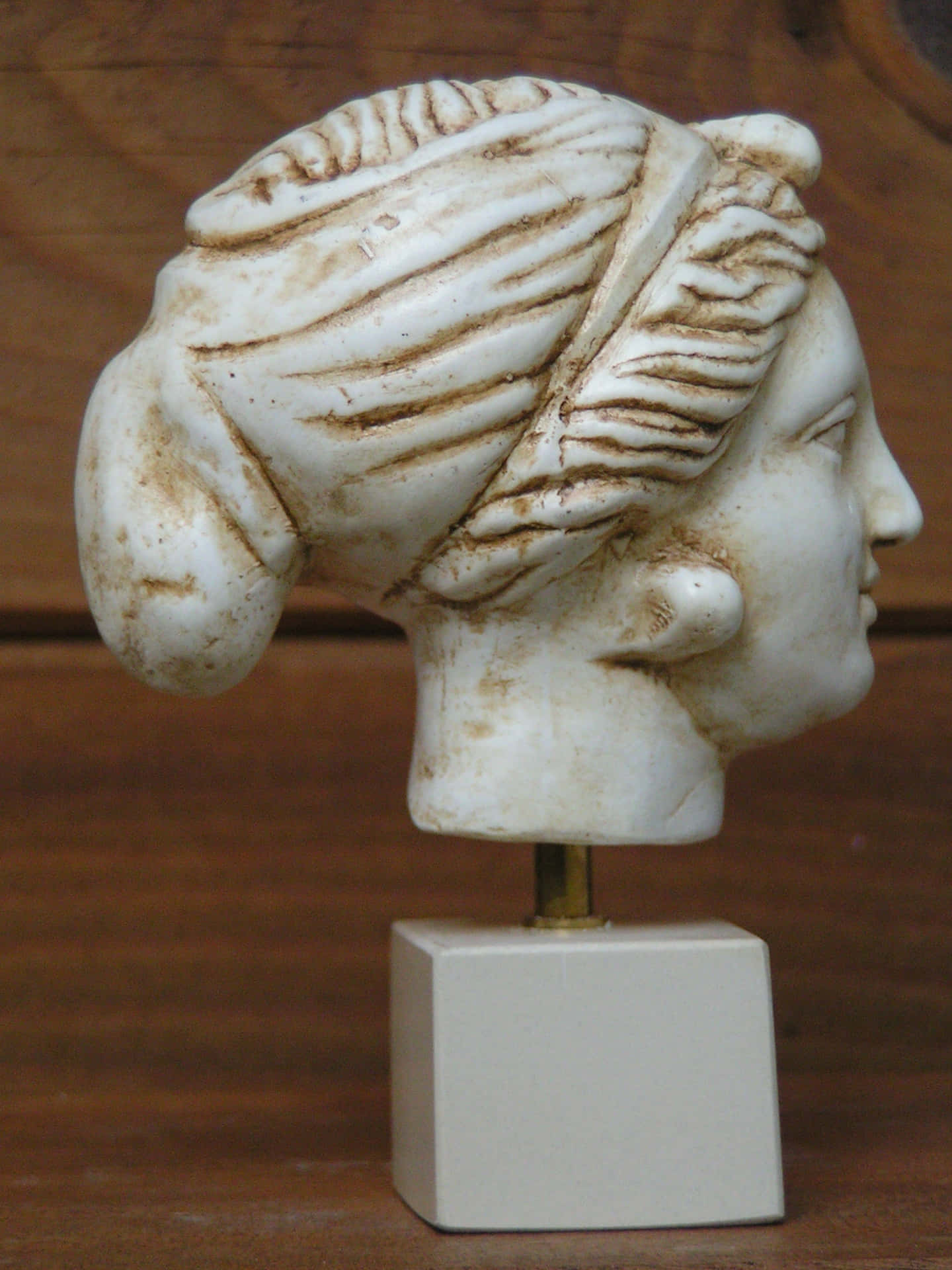  壁紙 — 古代大理石女性半身像，頭髮雕刻精細，坐在白色底座上，背景是溫暖的木質背景。