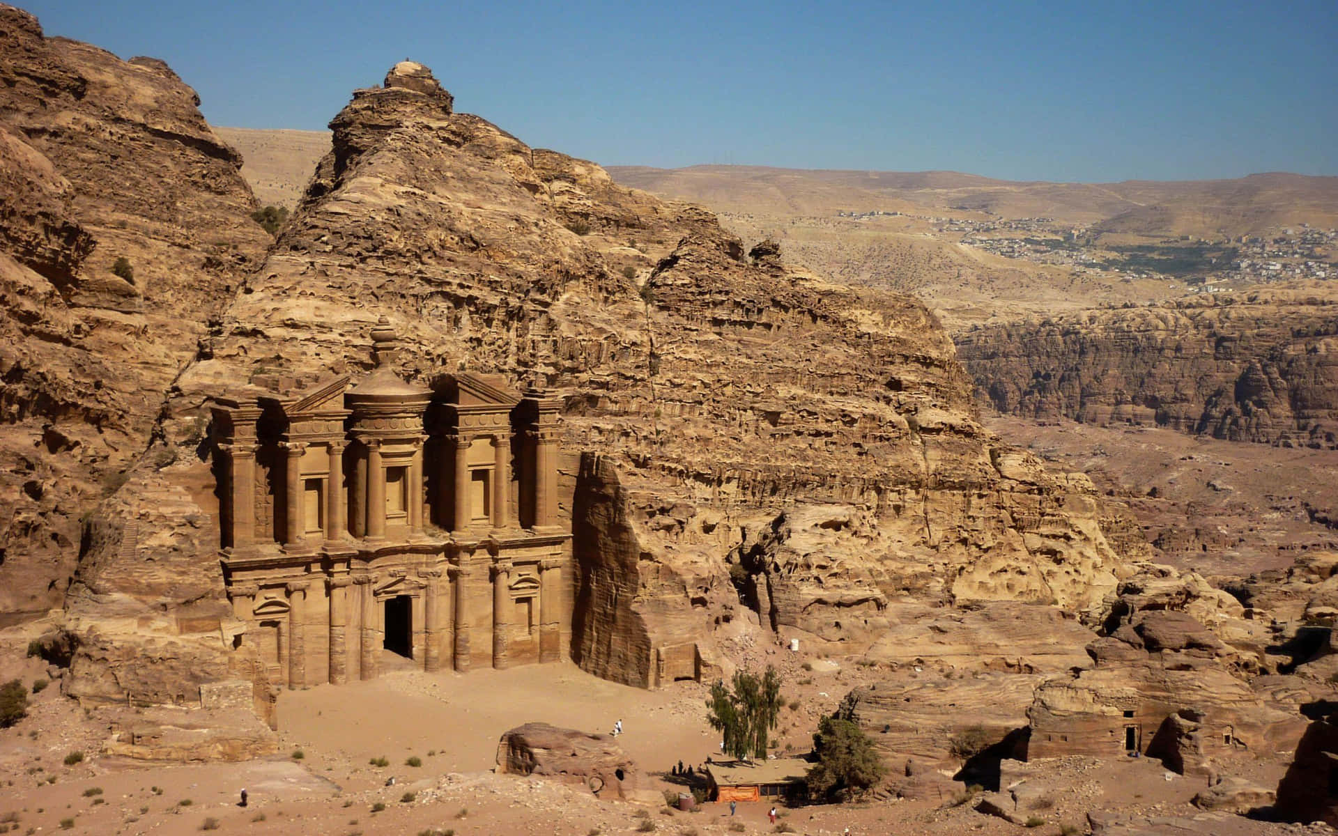 沙漠峽谷中雕刻的古老寺廟，藍天和遠處小鎮。 — Jordan 壁紙