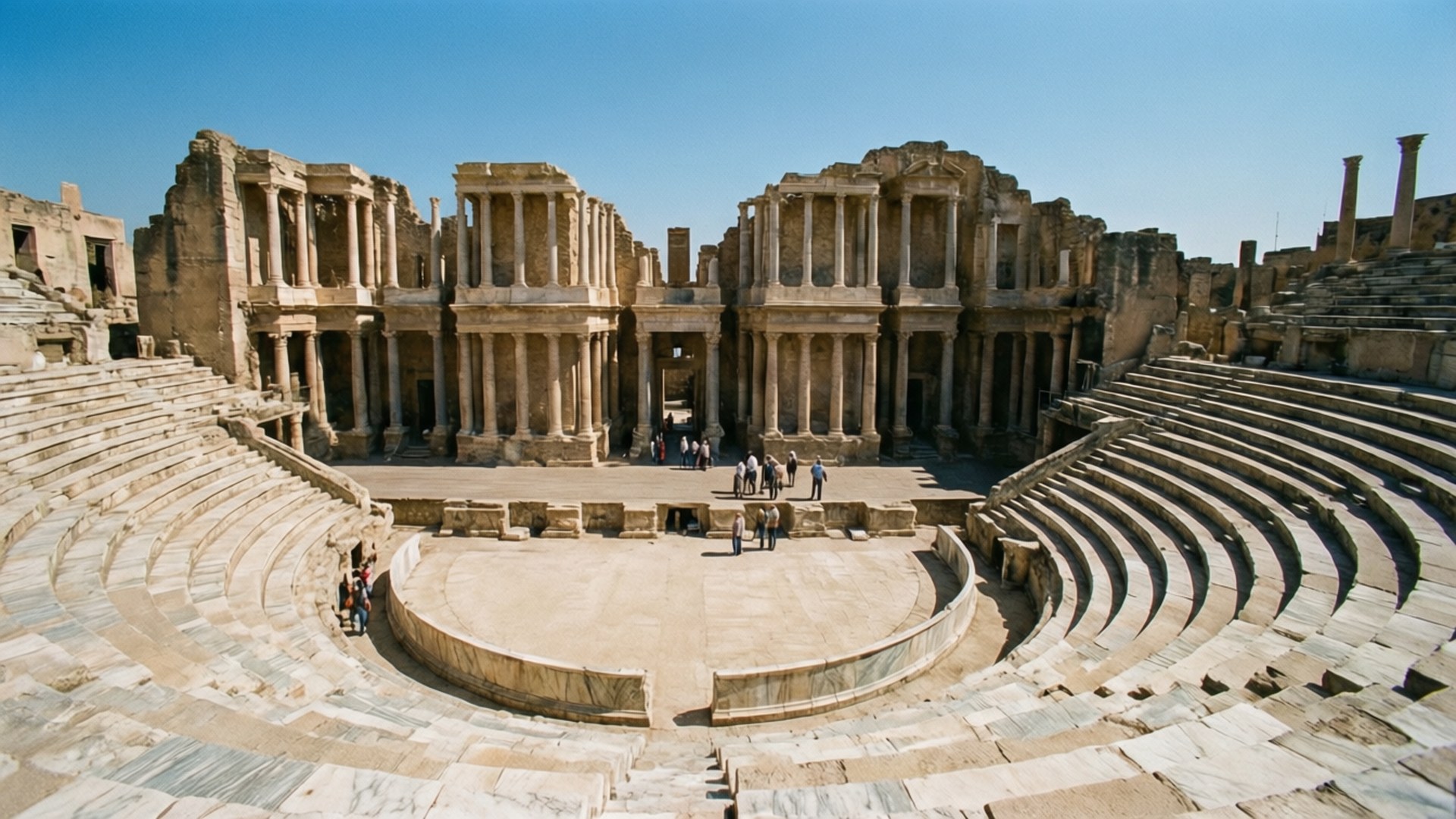 Roman Amphitheatre Ruins 壁紙 — 古老石造圓形劇場，擁有弧形分層座位和高聳的柱子，在清澈的藍天下。
