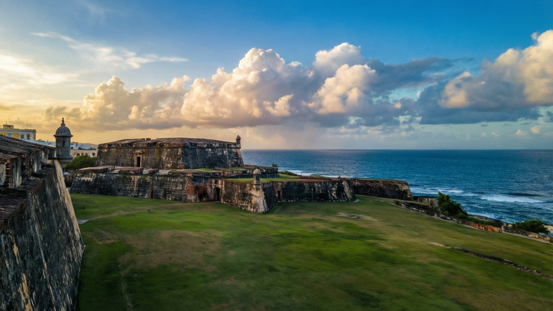 El Morro Sunset View 壁紙 — 古老石堡壘，草地庭院俯瞰碧綠海洋，天空有戲劇性的日落雲彩。