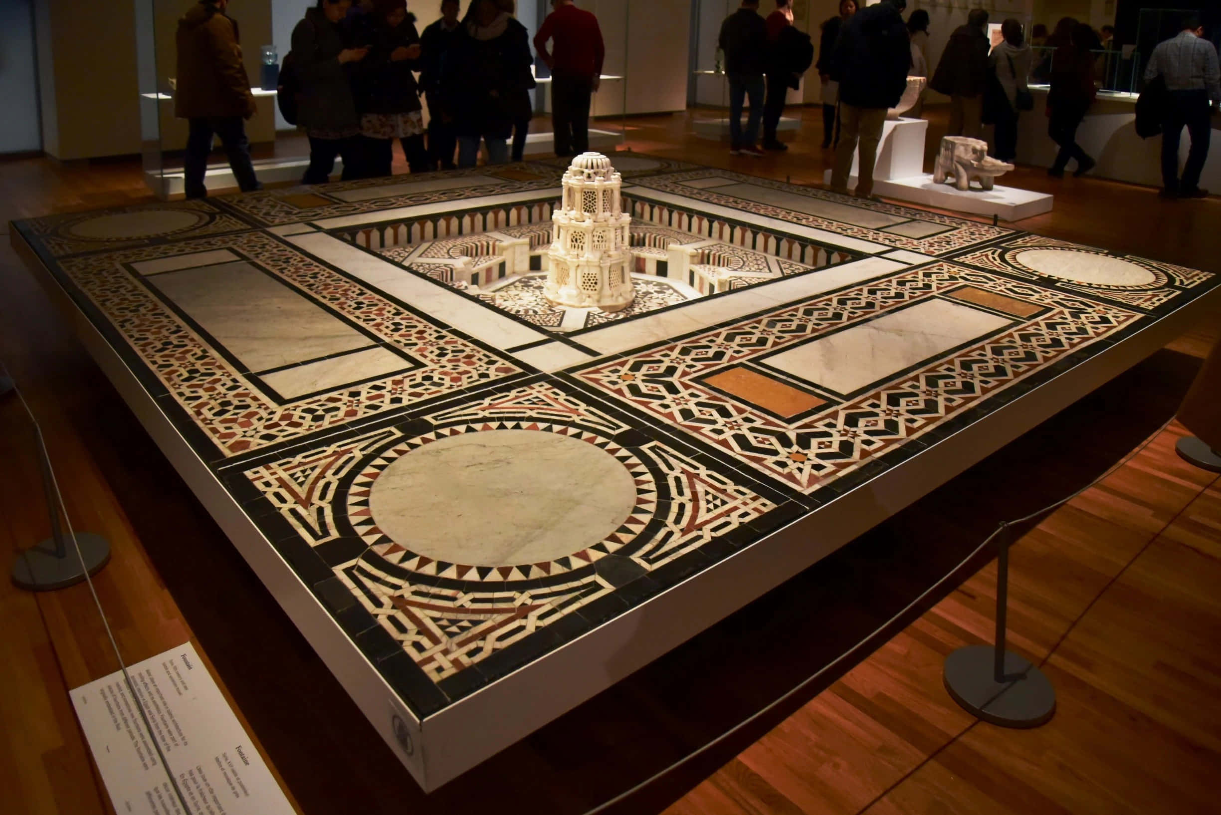 Aga Khan Museum 桌布 壁紙 — 一個詳細的圓頂建築模型放置在博物館畫廊的裝飾幾何馬賽克地板上，有參觀者在觀察。