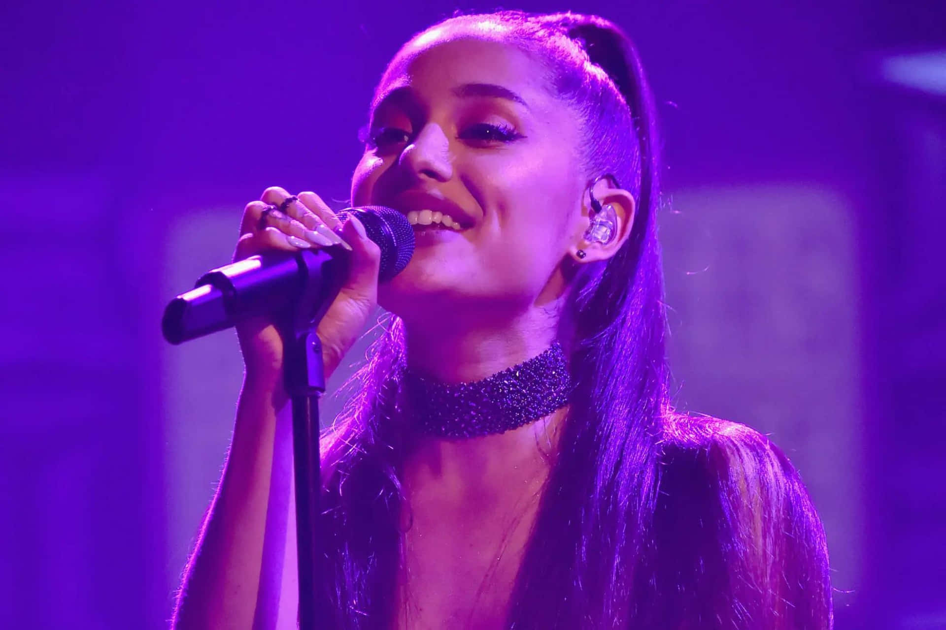  壁紙 — Ariana Grande 在充滿活力的紫色舞台燈光下對著麥克風唱歌，露出燦爛的笑容。