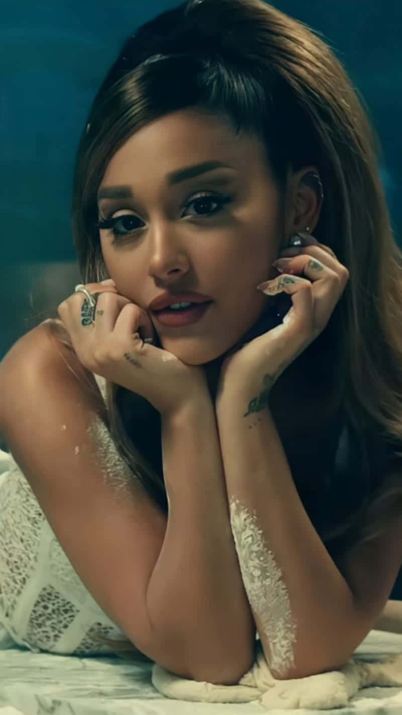  壁紙 — Ariana Grande 將下巴擱在紋身手上，身穿白色蕾絲上衣，背景為青綠色。