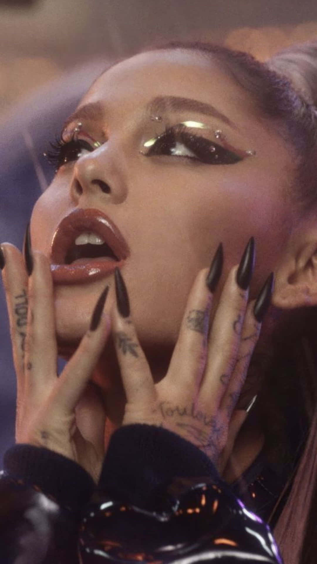  壁紙 — Ariana Grande 在舞台燈光下表演，帶有戲劇性的閃亮眼妝和長黑指甲，雙手框住她的臉。