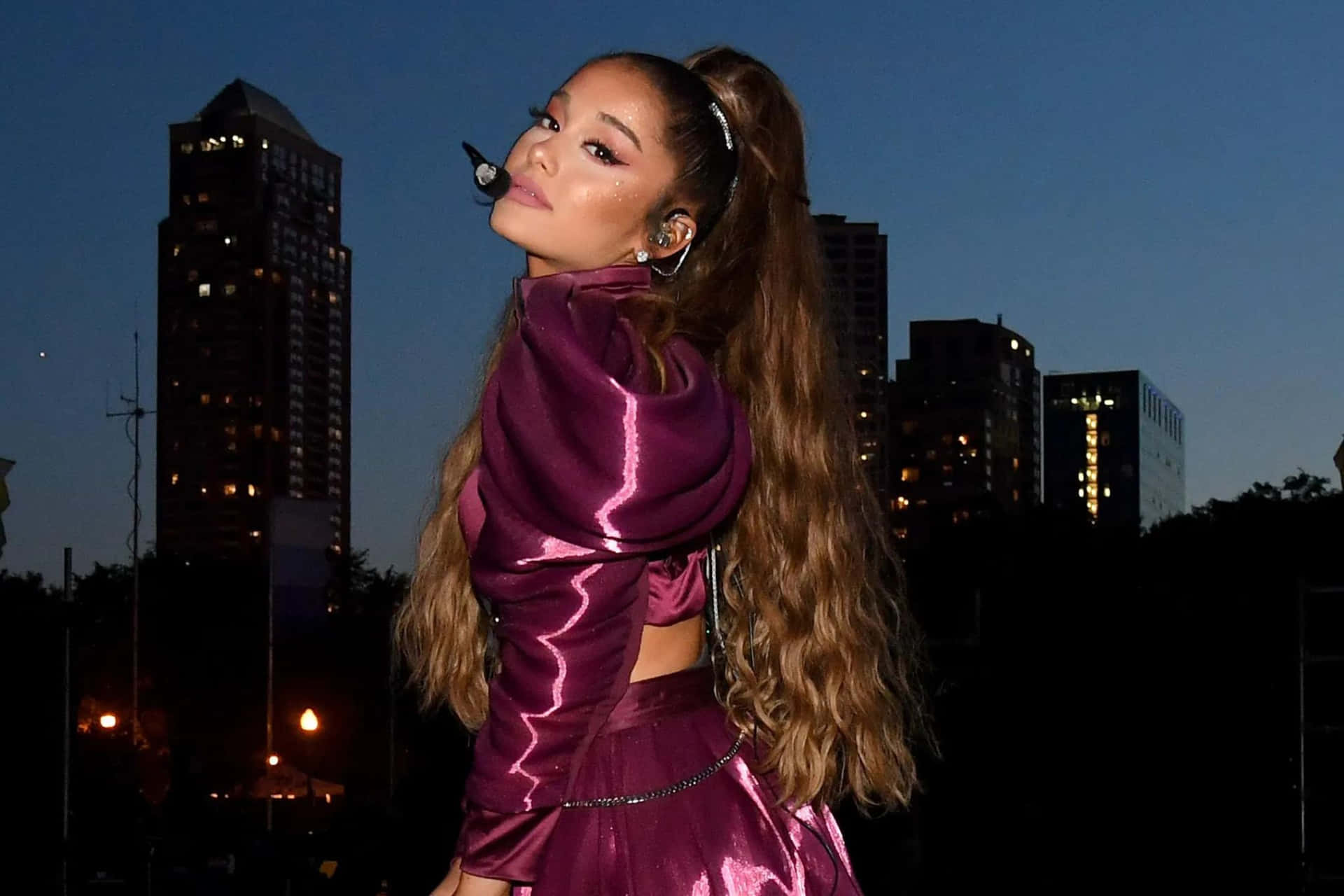  壁紙 — Ariana Grande 身穿閃亮紫色服裝，在暮色城市天際線前自信擺姿勢，身後是燈火通明的摩天大樓。