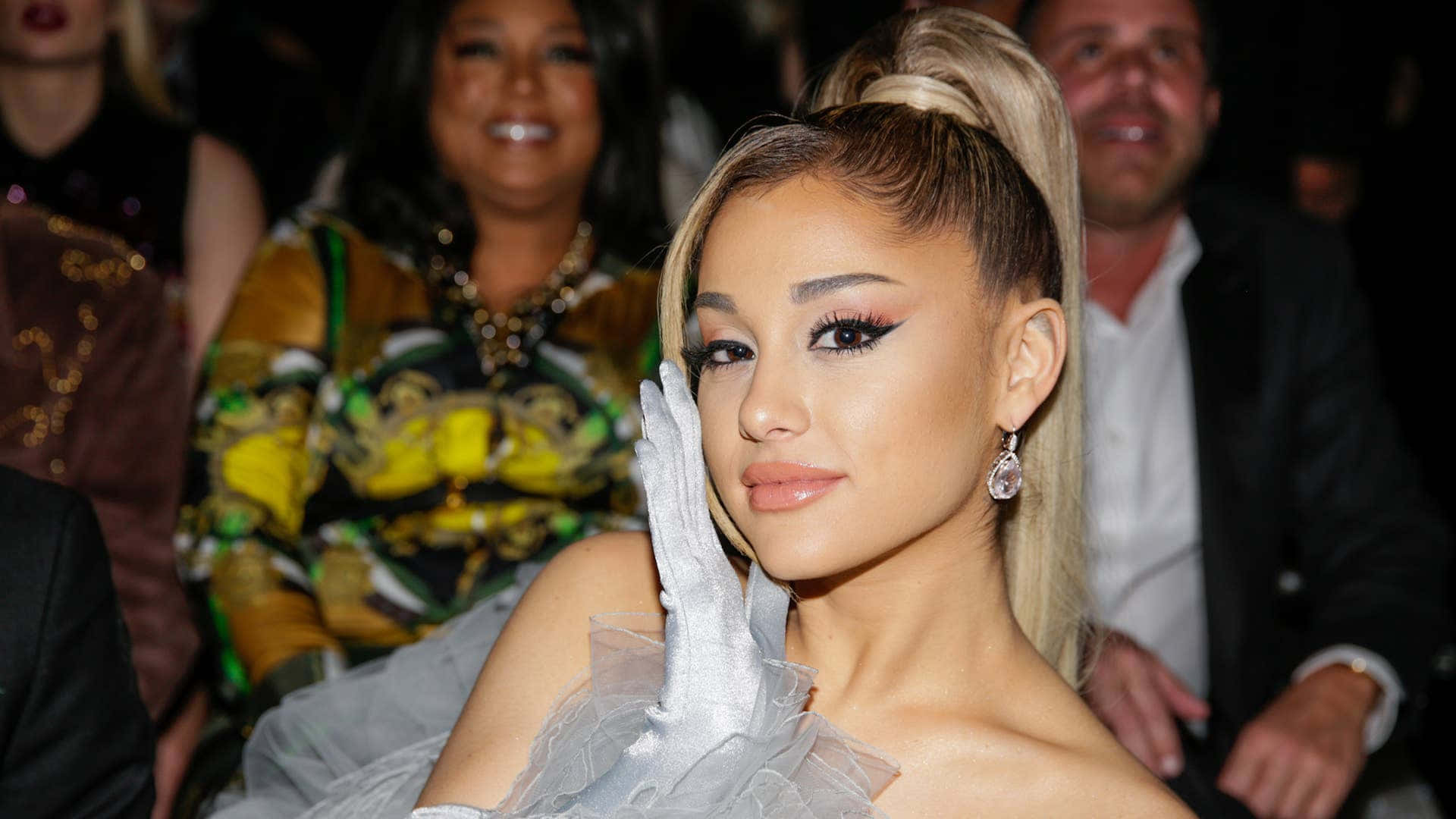  壁紙 — Ariana Grande 身穿銀色禮服和手套，在一個華麗的活動中優雅地擺姿勢，身後有模糊的與會者。