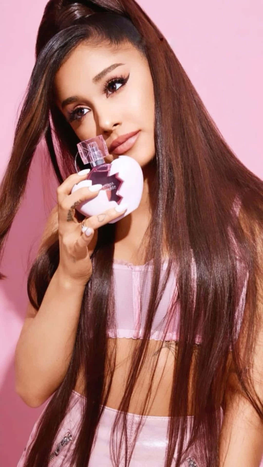  壁紙 — Ariana Grande 手持粉紅色香水瓶，背景為純粉紅色，長棕髮，妝容柔和。