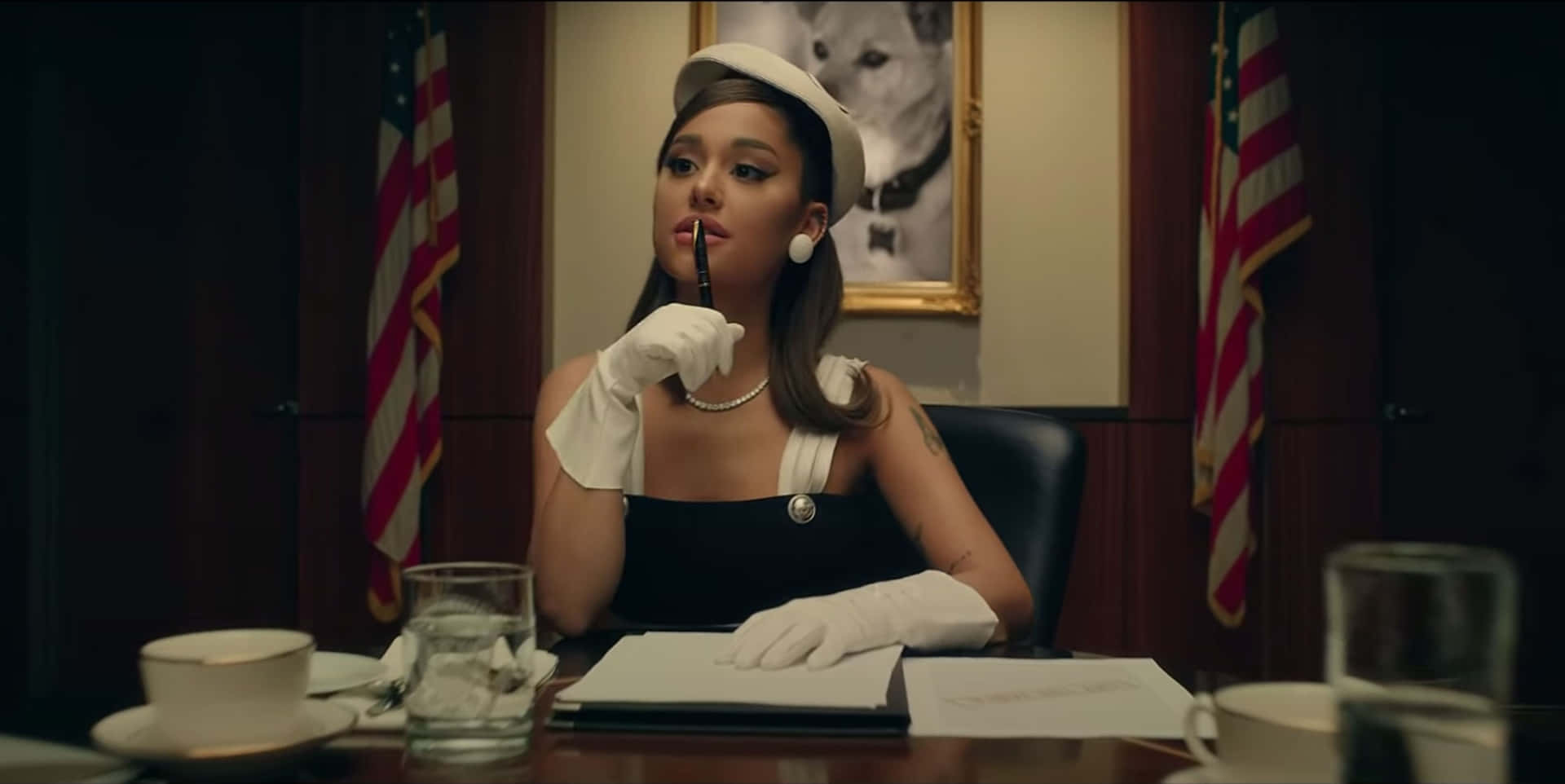  壁紙 — Ariana Grande 沉思地坐在一張木桌旁，身處一個燈光昏暗的辦公室，兩旁有美國國旗，戴著白色帽子和手套。
