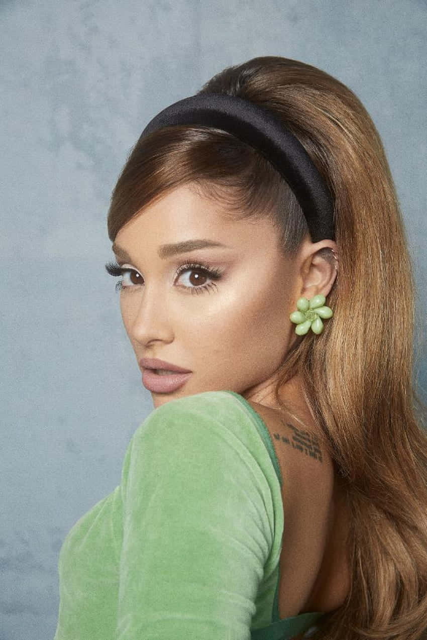 壁紙 — Ariana Grande 轉頭越過肩膀，身穿綠色天鵝絨上衣和花卉耳環，背景為帶紋理的藍灰色。