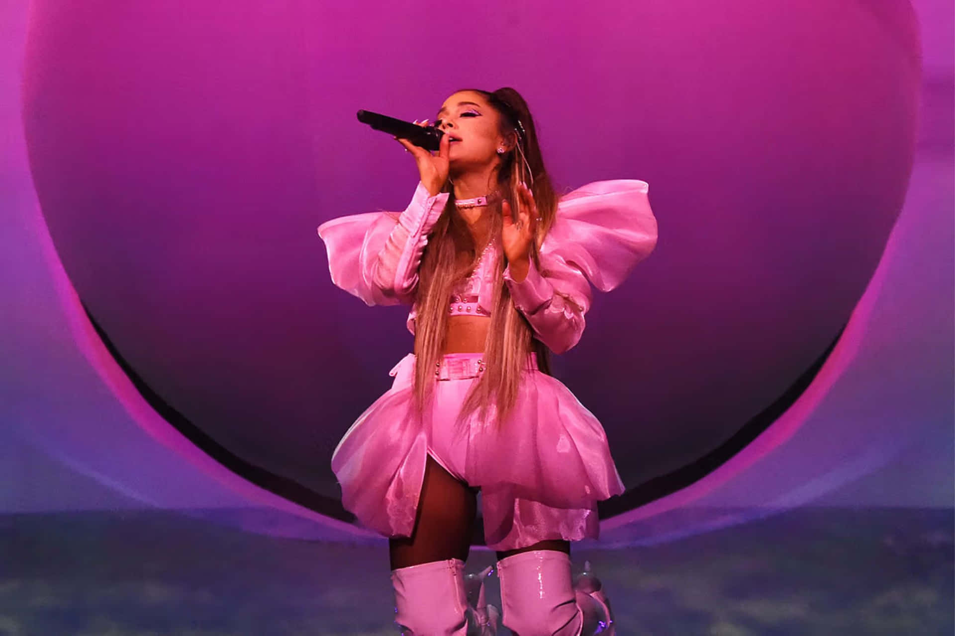  壁紙 — Ariana Grande 在充滿活力的紫色燈光下，穿著寬鬆的粉紅色服裝在舞台上表演。