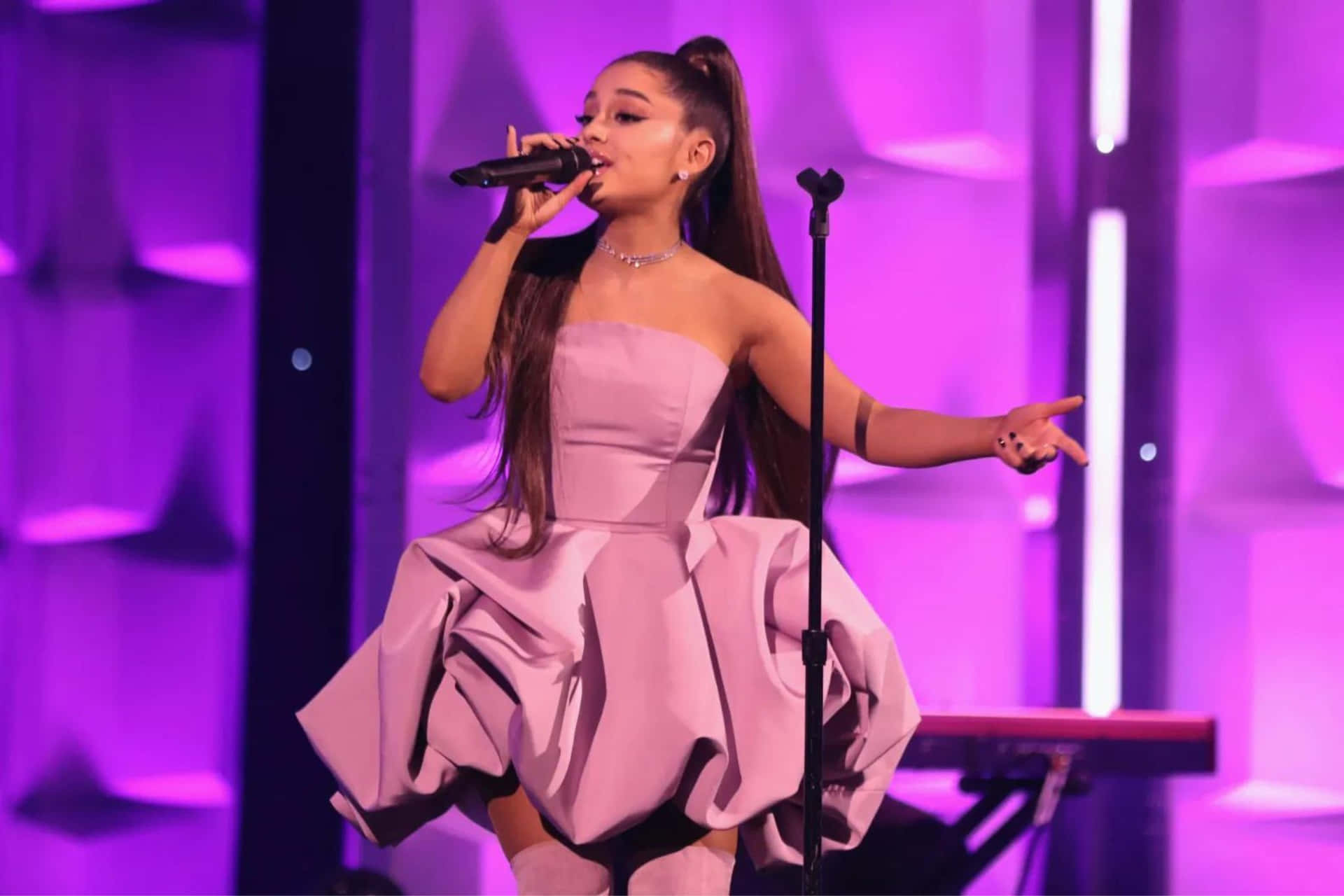  壁紙 — Ariana Grande 在充滿活力的紫色舞台燈光下，穿著飄逸的薰衣草色連衣裙在舞台上表演。