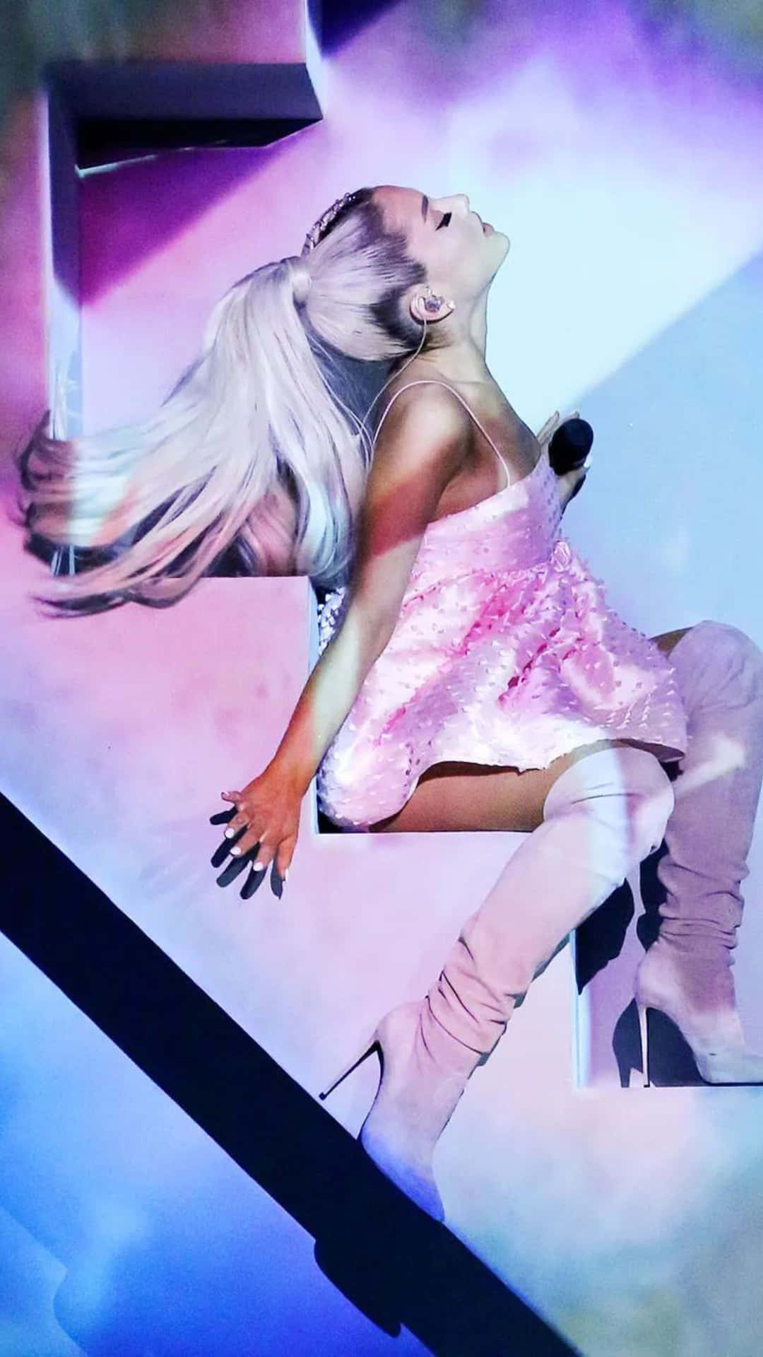  壁紙 — Ariana Grande 在舞台上表演，身穿閃亮粉紅色服裝，金色長髮飄逸，在充滿活力的紫色和藍色燈光下。
