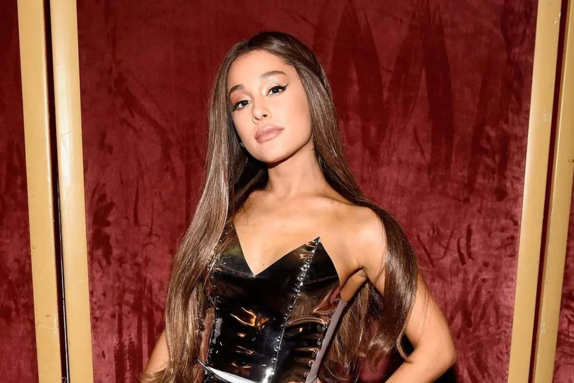  壁紙 — Ariana Grande 在一個有金色飾邊的濃郁紅色天鵝絨背景下，自信地擺出姿勢，穿著一件閃亮的黑色束胸。