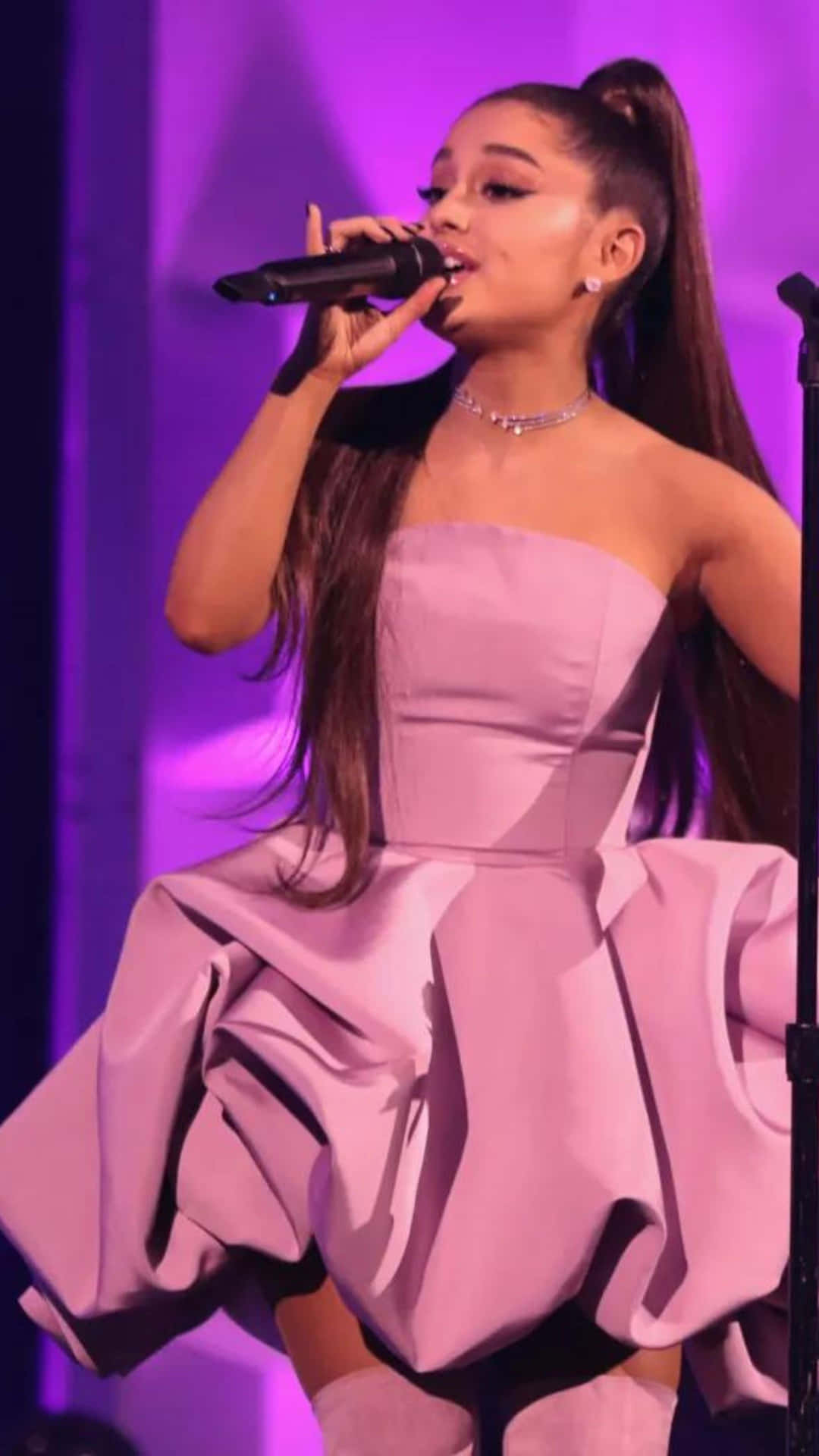  壁紙 — Ariana Grande 在紫色舞台燈光下，穿著寬鬆的薰衣草色長裙，熱情地對著麥克風唱歌。