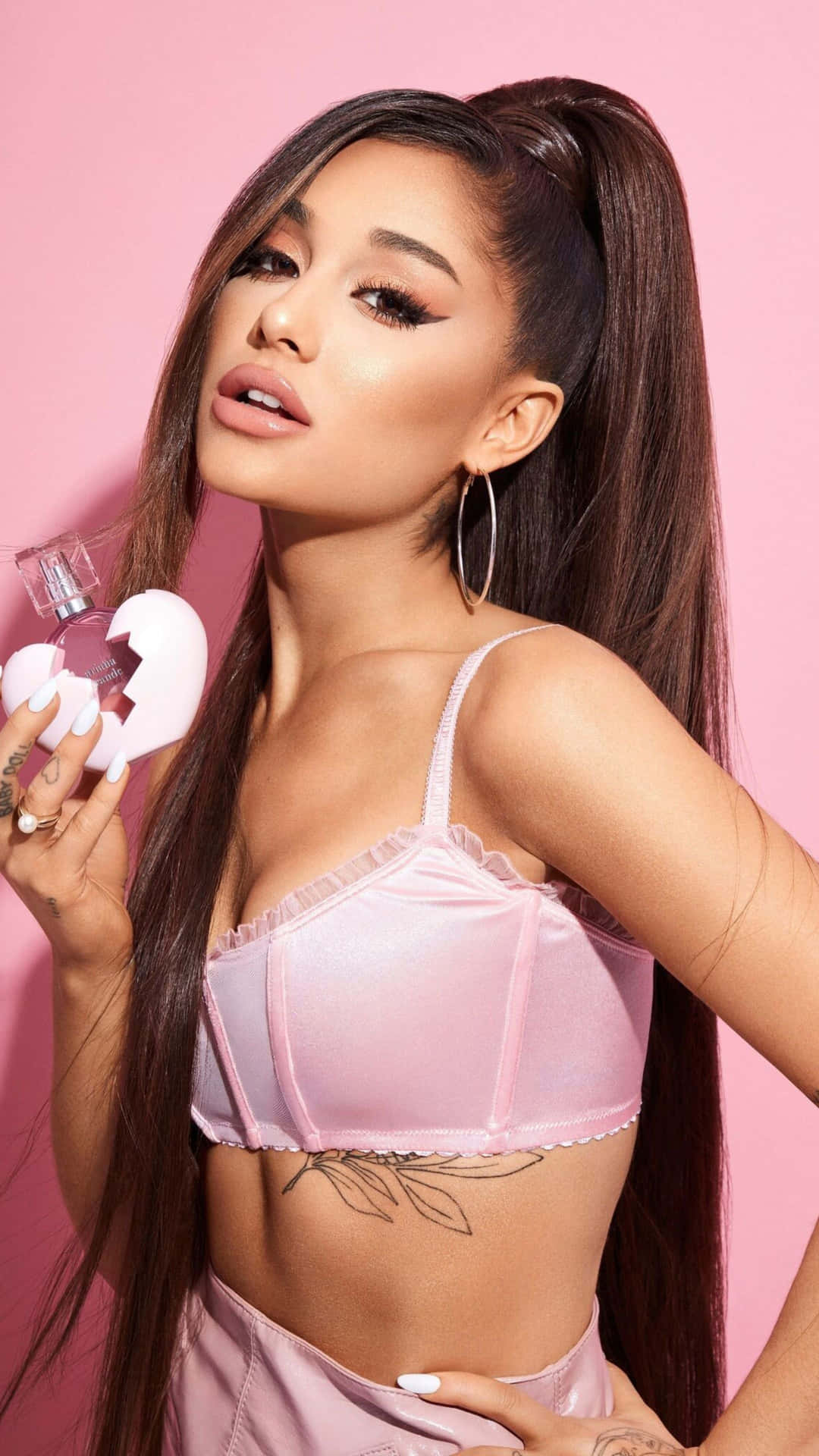  壁紙 — Ariana Grande 自信地穿著粉紅色內衣，手持一個破碎心形香水瓶，背景為純粉紅色。