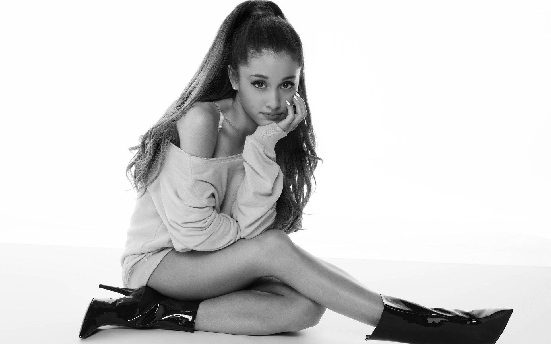 Grande 桌布 壁紙 — Ariana Grande 優雅地坐在單色工作室照片中，手托著臉頰，穿著寬鬆毛衣和亮面靴子。