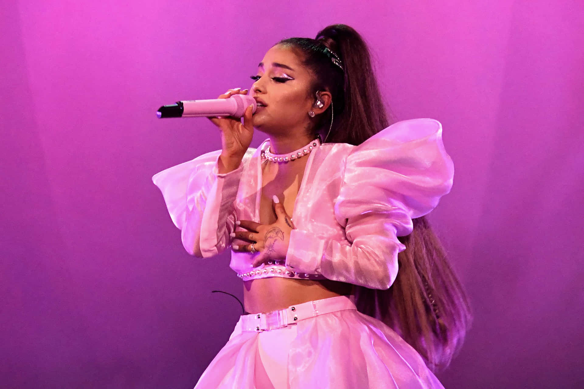 亞莉安娜·格蘭德 桌布 壁紙 — Ariana Grande 在充滿活力的紫色舞台背景下，穿著閃閃發光的粉紅色服裝熱情表演。