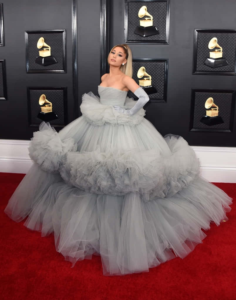 亞莉安娜·格蘭德 桌布 壁紙 — Ariana Grande 在紅地毯上，身穿灰色薄紗長裙，背景是黑色牆壁，上面裝飾著葛萊美獎座。
