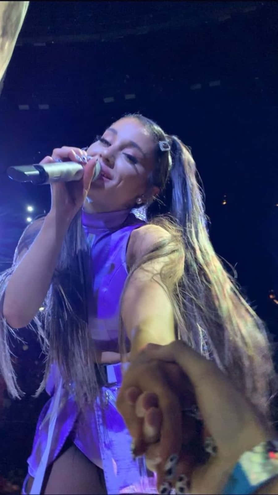 Aesthetic Ariana Grande 桌布 壁紙 — Ariana Grande 在紫色燈光下於舞台上對著麥克風唱歌，同時在前景中握著一位粉絲的手。