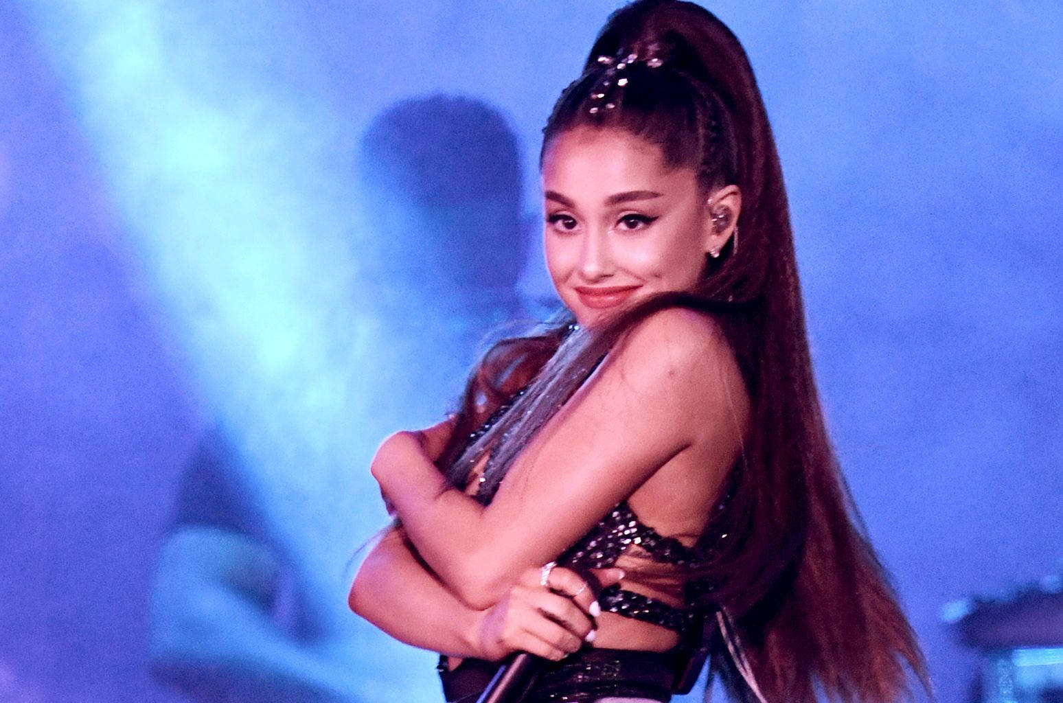 高清 桌布 壁紙 — Ariana Grande 在藍色舞台燈光下，身穿閃亮黑色服裝，交叉雙臂，在舞台上溫暖地微笑。