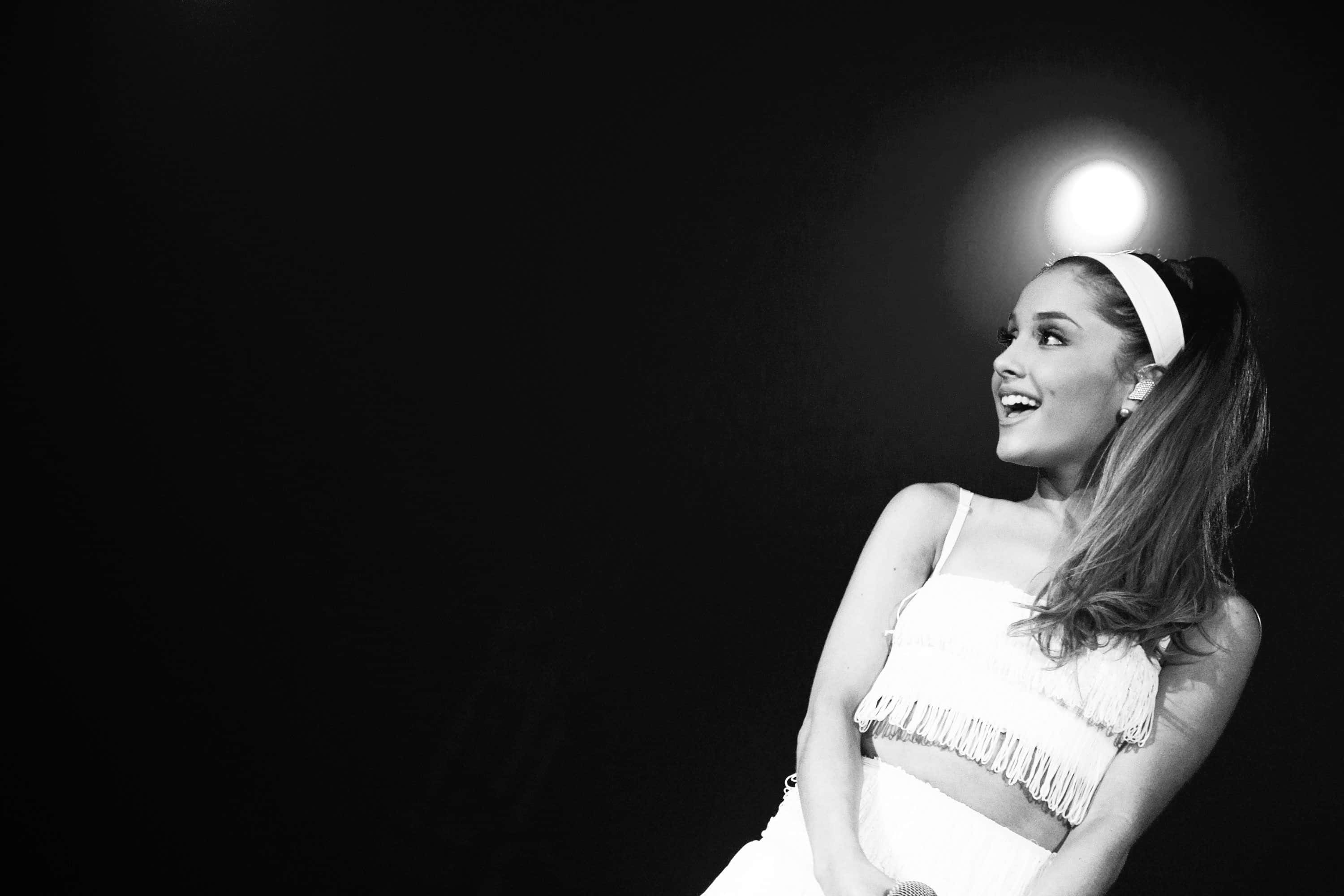 Ariana Grande Aesthetic 桌布 壁紙 — Ariana Grande 在黑暗舞台背景下，聚光燈下穿著白色流蘇服裝，燦爛微笑。