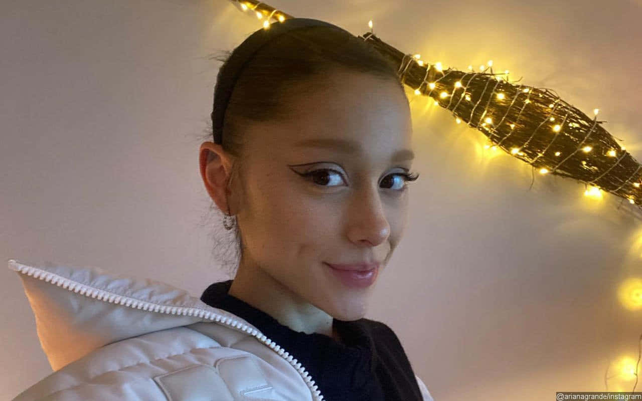 亞莉安娜·格蘭德 桌布 壁紙 — Ariana Grande 穿著白色羽絨外套，頭頂有溫暖的仙女燈和裝飾樹枝，溫柔地微笑。