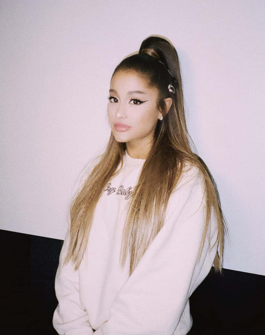 亞莉安娜·格蘭德 桌布 壁紙 — Ariana Grande 穿著柔軟的白色衛衣，頭髮紮成高馬尾，靠著素牆擺姿勢。