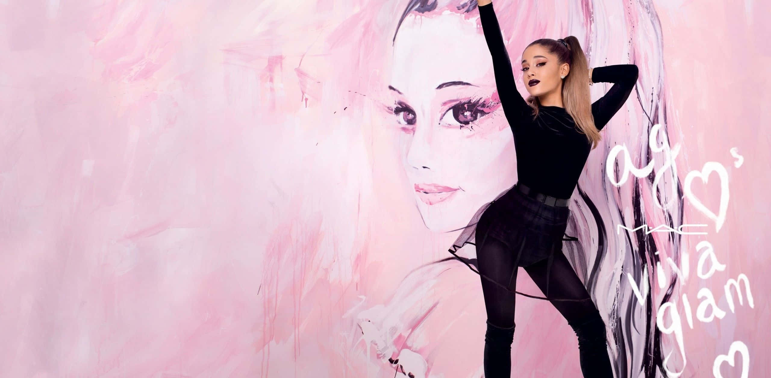 Aesthetic Ariana Grande 桌布 壁紙 — Ariana Grande 自信地站在一幅她臉部的大型粉紅色彩繪壁畫前，帶有 MAC Viva Glam 品牌標誌。