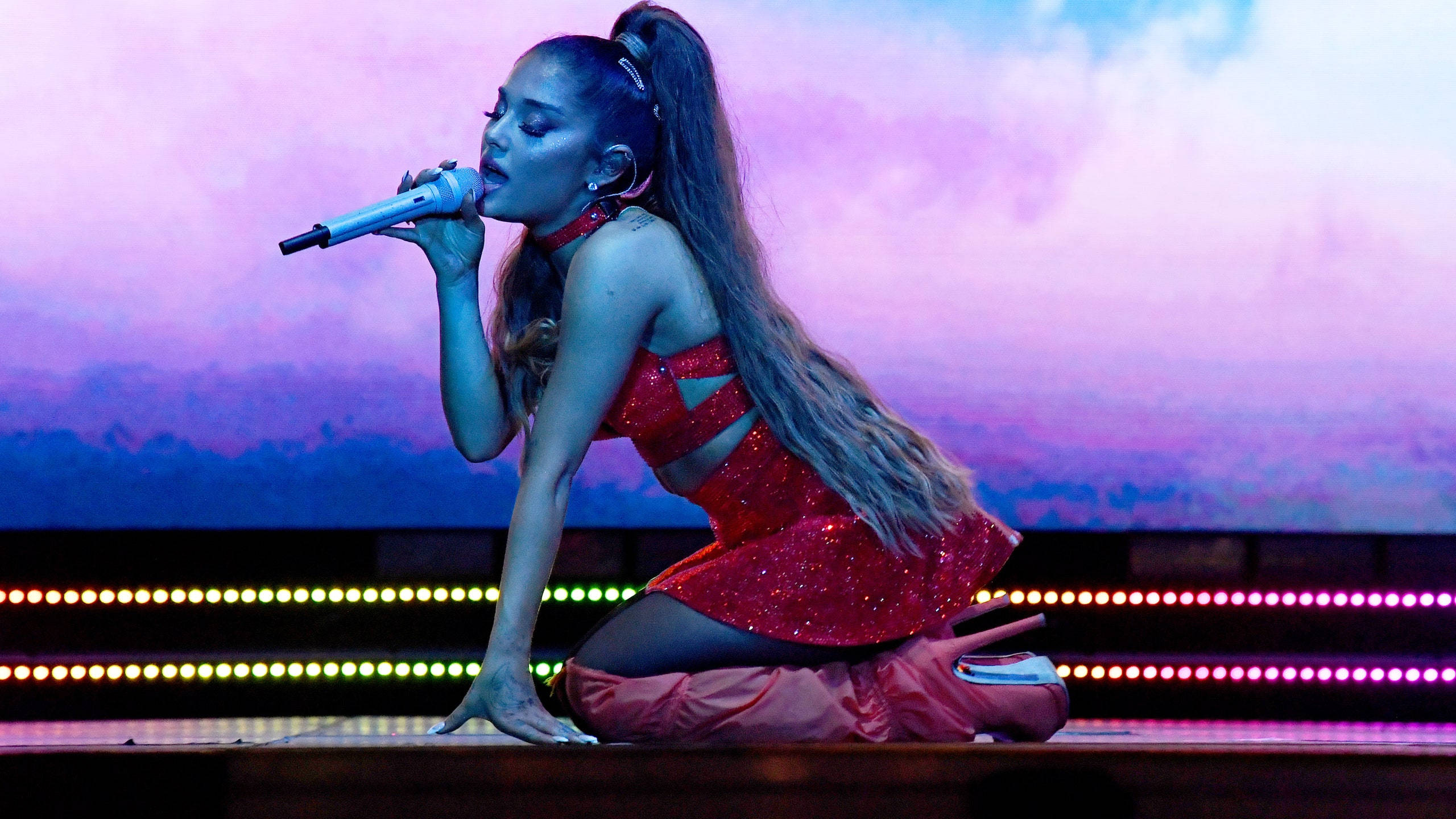 Lollapalooza 桌布 壁紙 — Ariana Grande 跪在舞台上，身穿閃閃發光的紅色服裝，在充滿活力的紫色和粉紅色燈光下對著麥克風唱歌。
