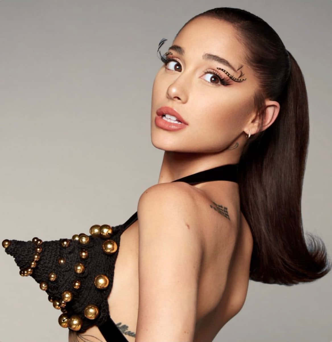 亞莉安娜·格蘭德 桌布 壁紙 — Ariana Grande 身穿黑色珠飾上衣，帶有金色點綴，搭配戲劇性的翼狀睫毛，在灰色中性背景前擺姿勢。