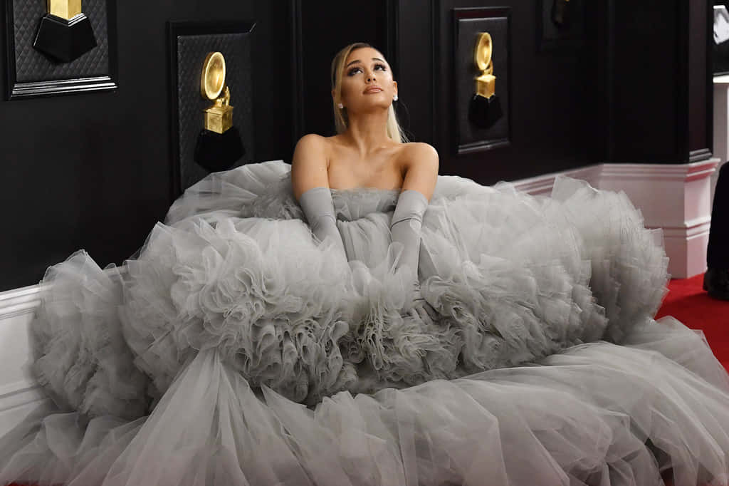 亞莉安娜·格蘭德 桌布 壁紙 — Ariana Grande 身穿寬大的灰色薄紗禮服，端莊地坐在紅地毯上，背景是裝飾著金色葛萊美獎項的黑色牆壁。