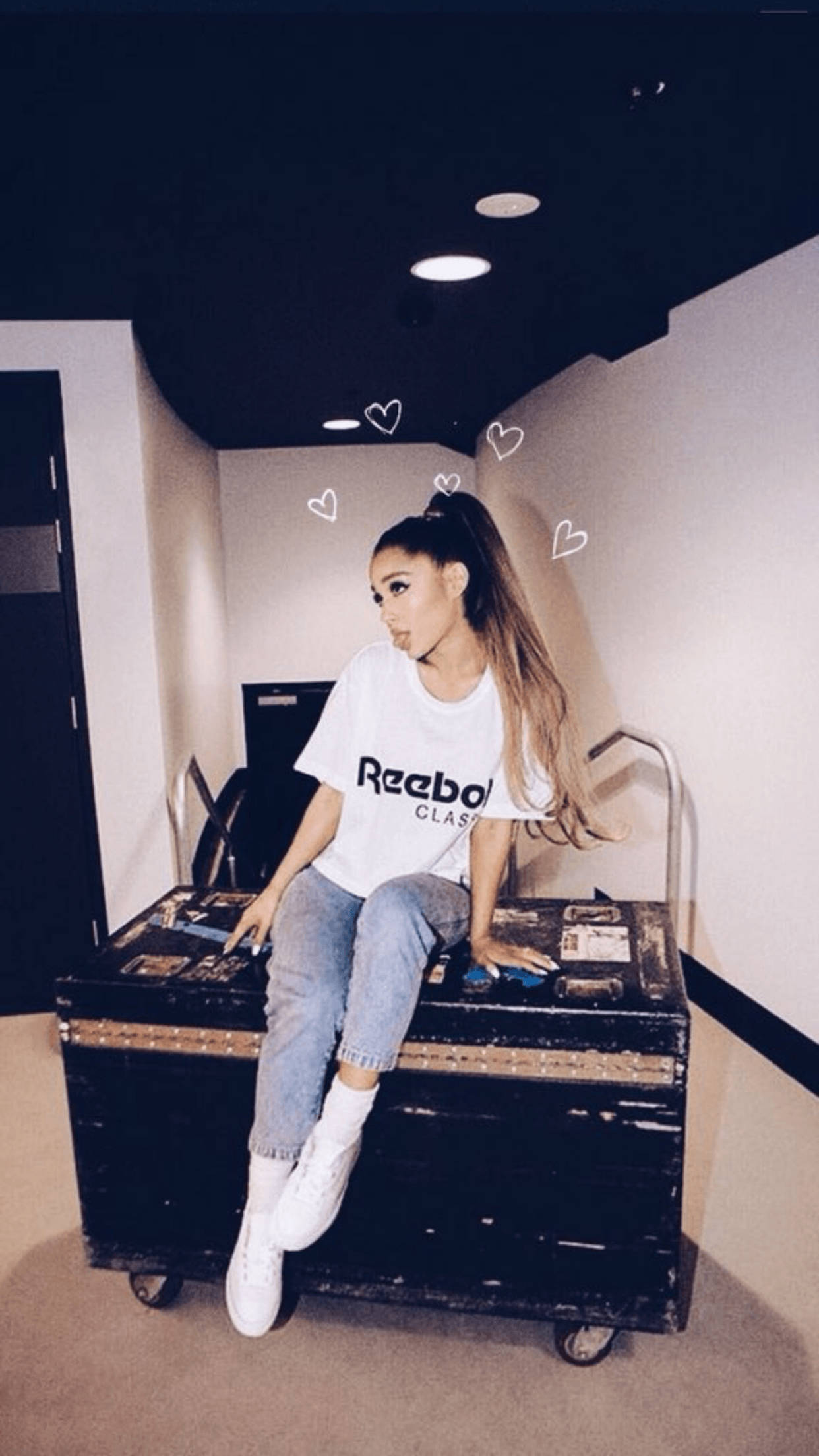 高清 桌布 壁紙 — Ariana Grande 隨意地坐在一個黑色有輪行李箱上，身處簡約風格的房間，頭頂上方有漂浮的心形塗鴉。