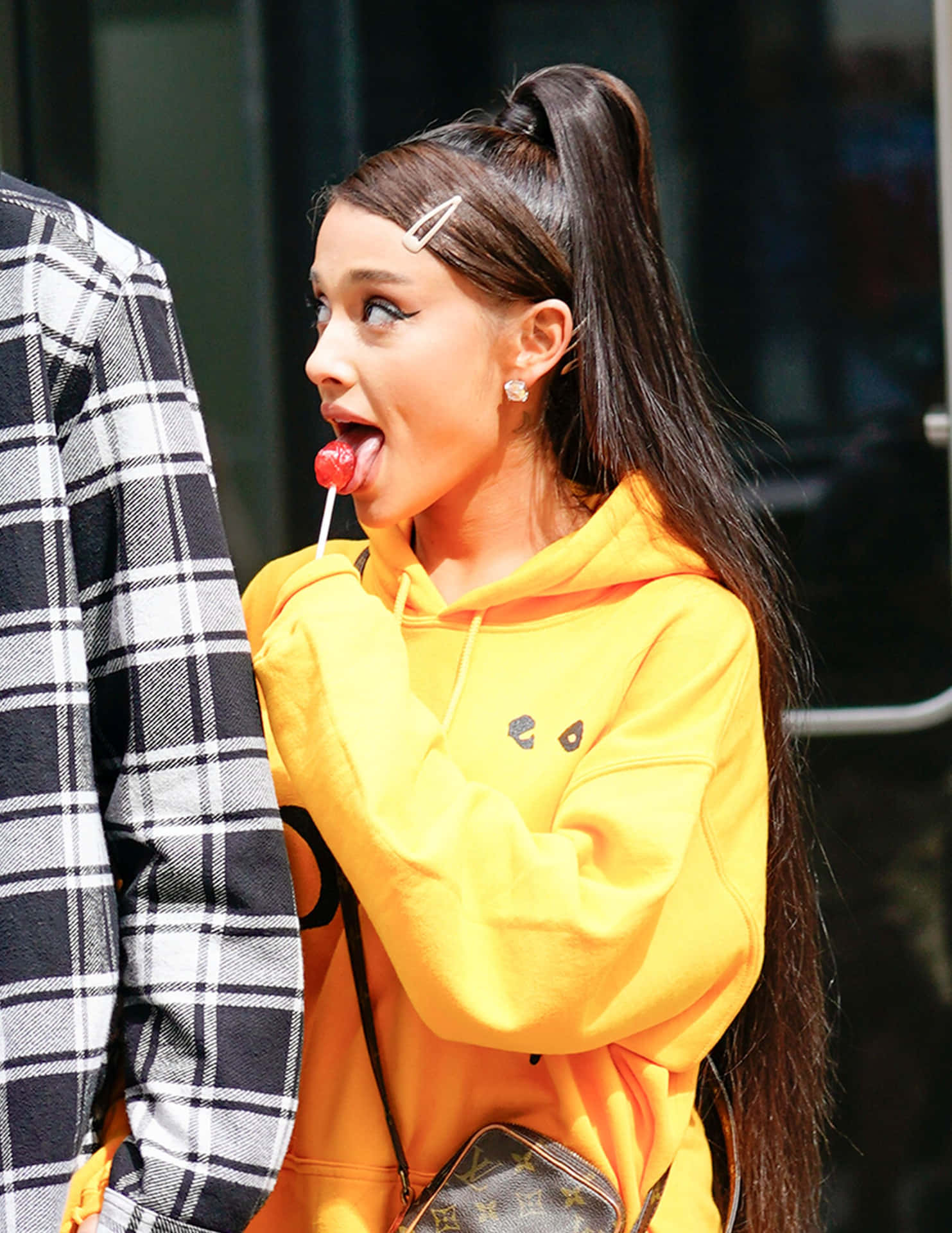亞莉安娜·格蘭德 桌布 壁紙 — Ariana Grande 頑皮地伸出舌頭，同時拿著紅色棒棒糖，身穿亮黃色連帽衫。
