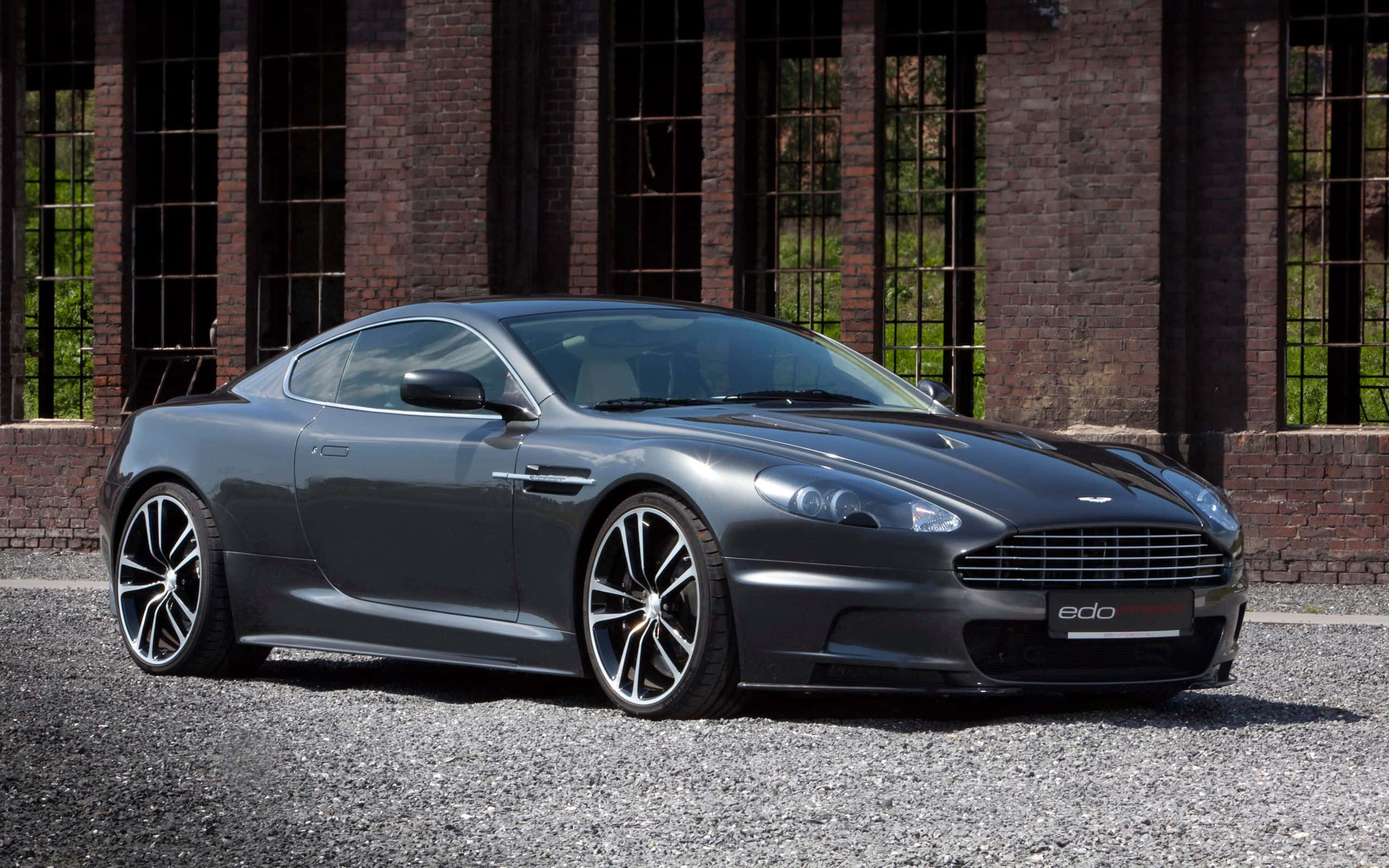 賽道上驚豔的銀色 Aston Martin DB9 壁紙 — 一架時尚啞灰色Aston Martin跑車停泊在碎石路上，前方是帶有高窗的風化磚建築。