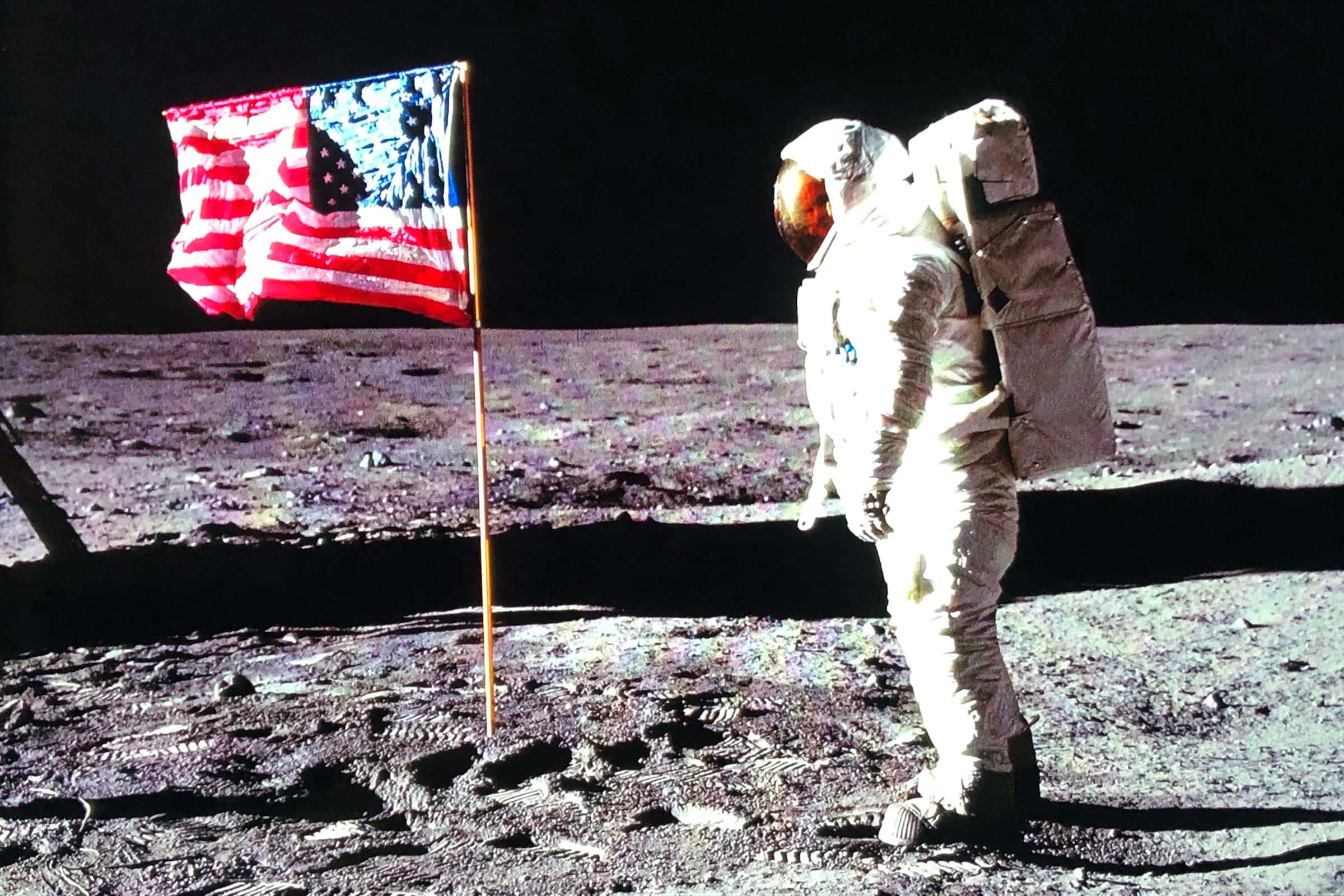 Astronaut On Moon 桌布 壁紙 — 一名太空人站在月球灰色的表面上，旁邊是一面在黑色天空下飄揚的美國國旗。