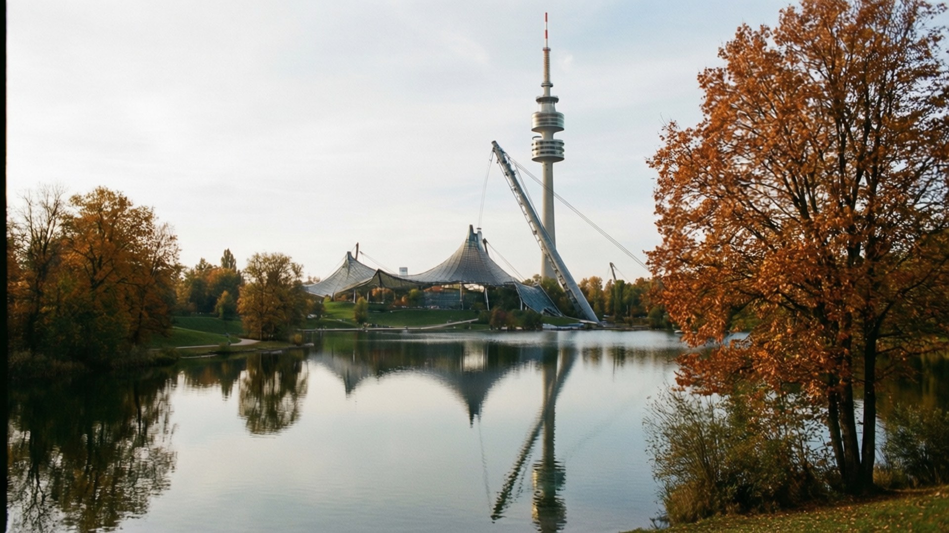 Olympiapark Autumn Mood 壁紙 — 秋天的樹木環繞著平靜的湖泊，倒映出標誌性的奧運塔和帳篷狀結構，天空多雲。
