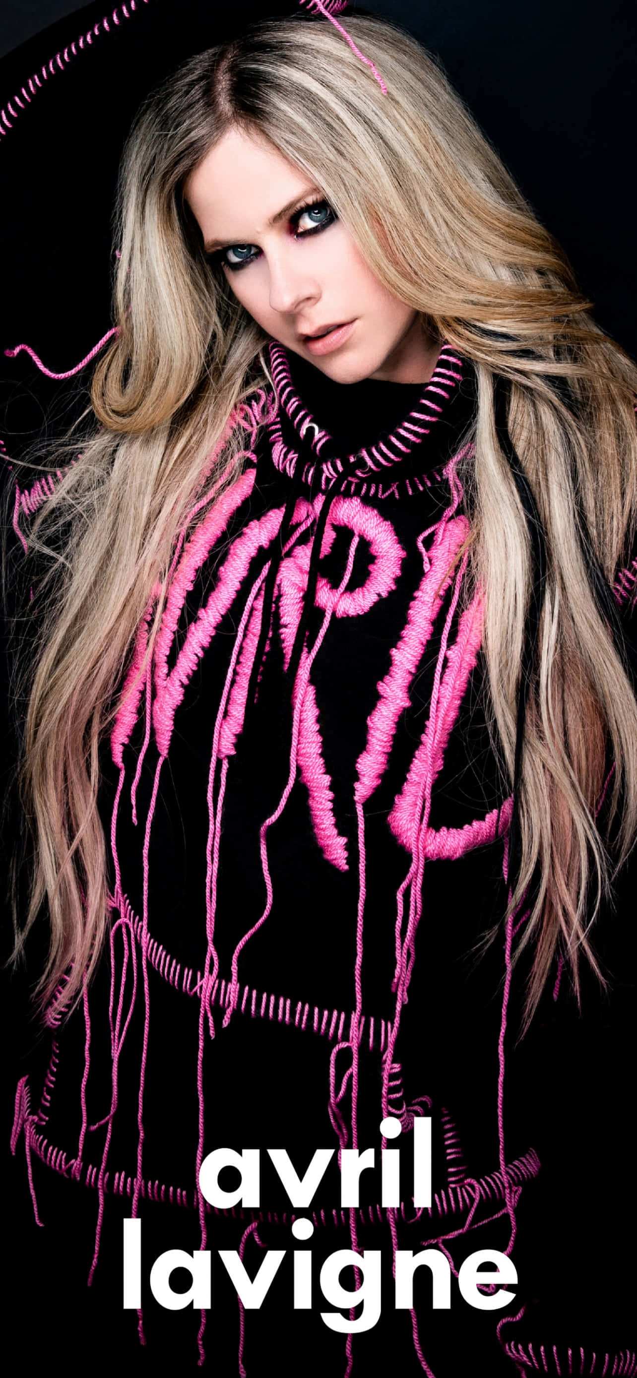 艾薇兒·拉維尼 桌布 壁紙 — Avril Lavigne 穿著黑色高領毛衣，帶有醒目的粉紅色縫線字母和流蘇，在黑暗背景下凝視。