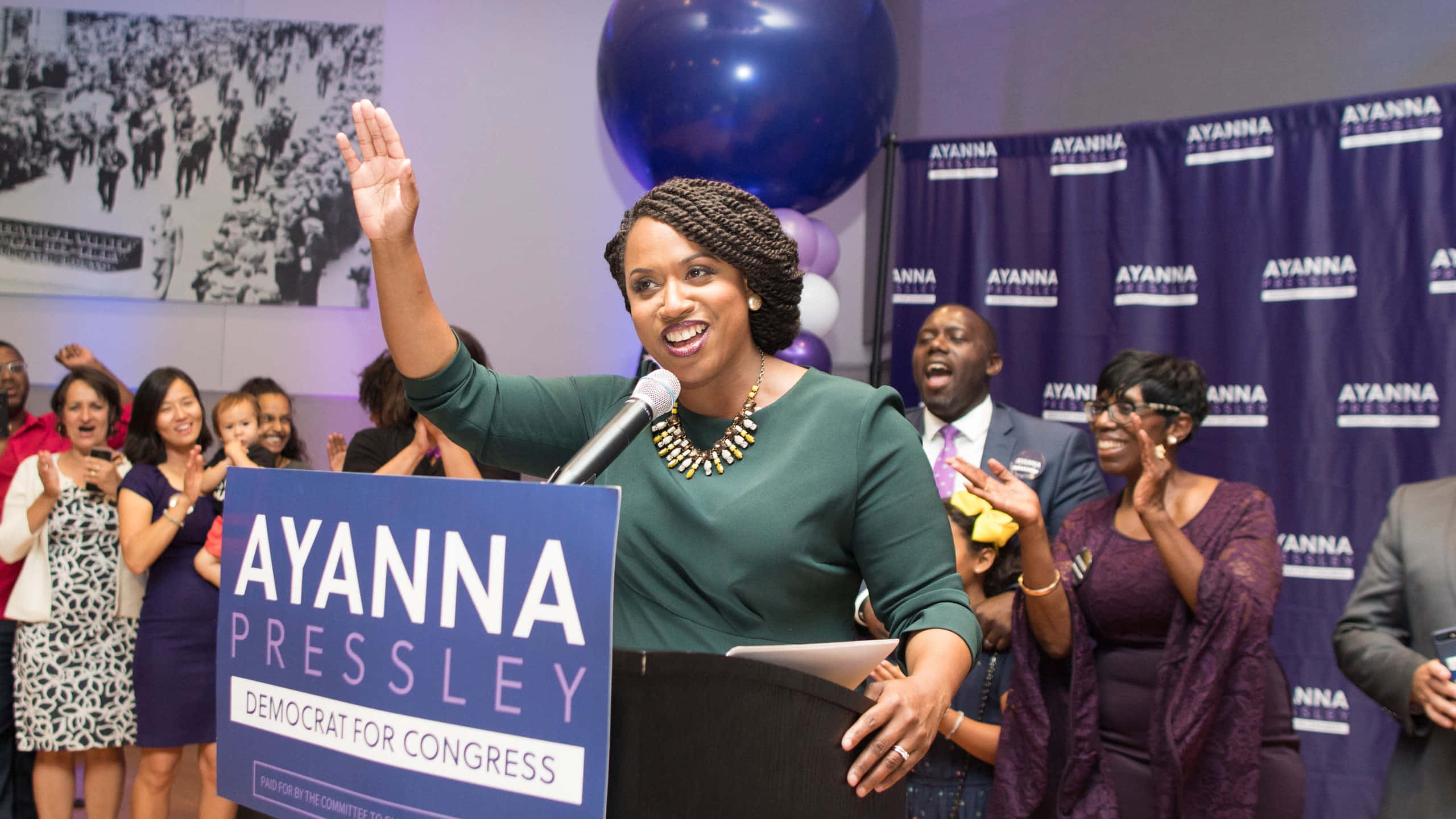 Public 桌布 壁紙 — Ayanna Pressley 在競選活動的講台上熱情揮手，支持者在身後歡呼。