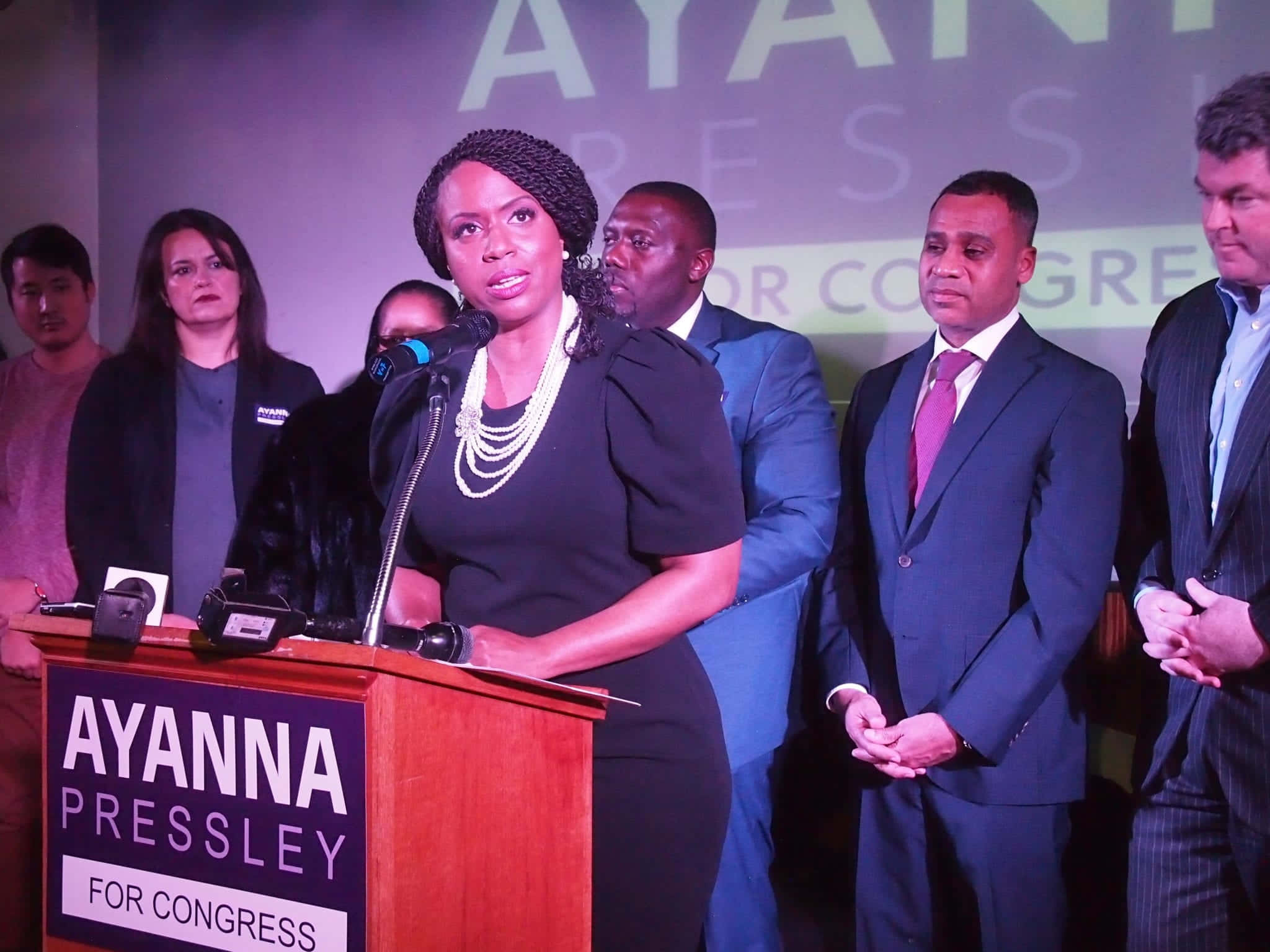 人物 桌布 壁紙 — Ayanna Pressley 在舞台燈光下於競選講台上發言，支持者站在她身後。