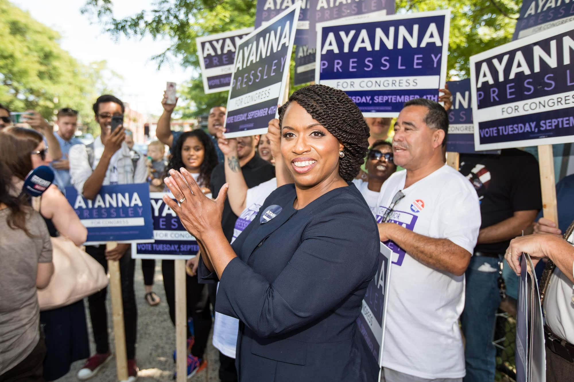 樹木 桌布 壁紙 — Ayanna Pressley 在陽光明媚的戶外競選集會上微笑鼓掌，周圍是舉著她標語牌的支持者。