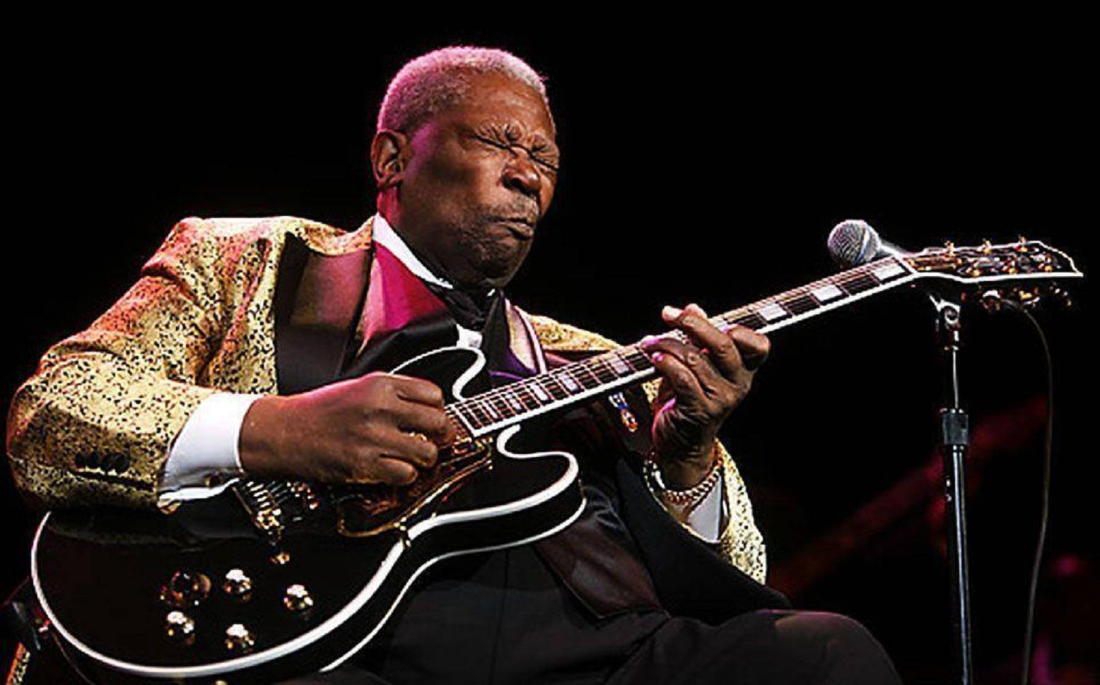 Gibson 桌布 壁紙 — B.B. King 在戲劇性的舞台燈光下，穿著閃閃發光的金色外套，熱情地彈奏他的黑色電吉他。