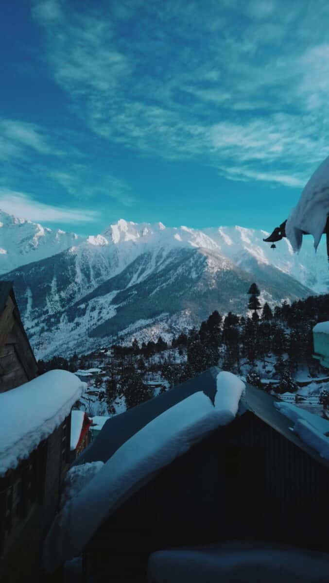  壁紙 — 白雪覆蓋的山脈在一個屋頂積雪的村莊後方升起，天空湛藍，雲彩縷縷。